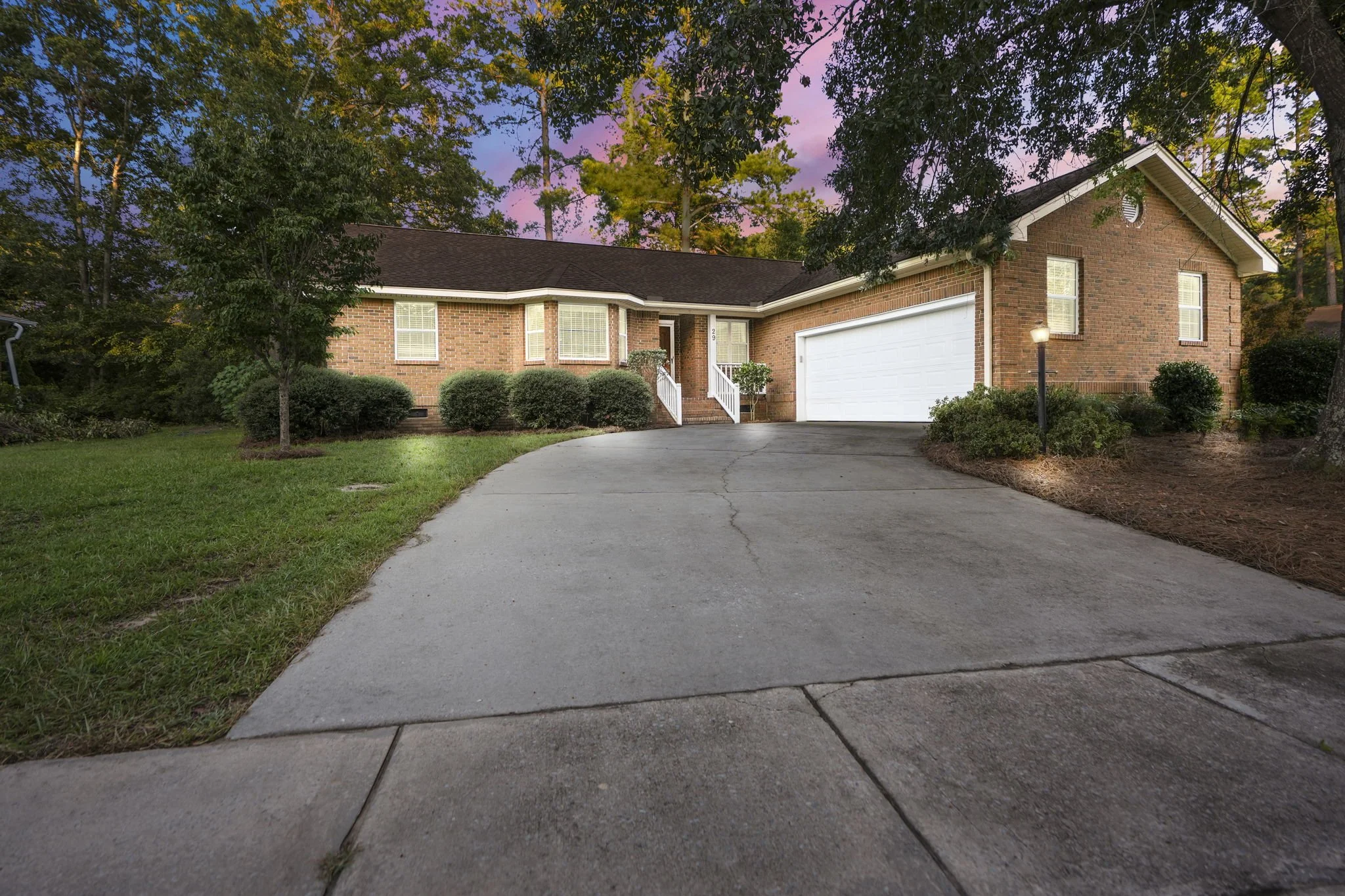 A brick house with a white garage door, front porch with small stairs, and surrounded by green bushes and trees during early evening with a colorful sky.