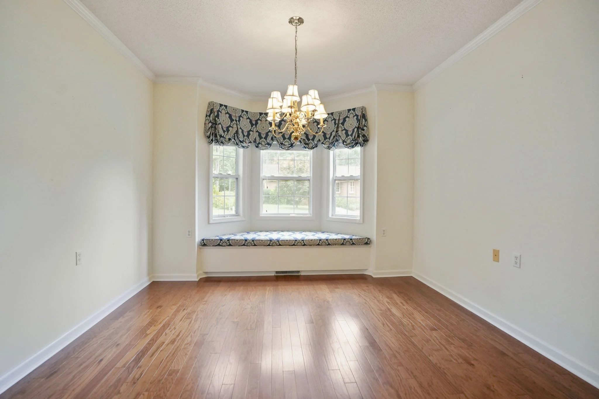 Empty room with hardwood floors, cream-colored walls, a bay window with a decorative valance and window seat, a chandelier hanging from the ceiling, and electrical outlets on the walls.
