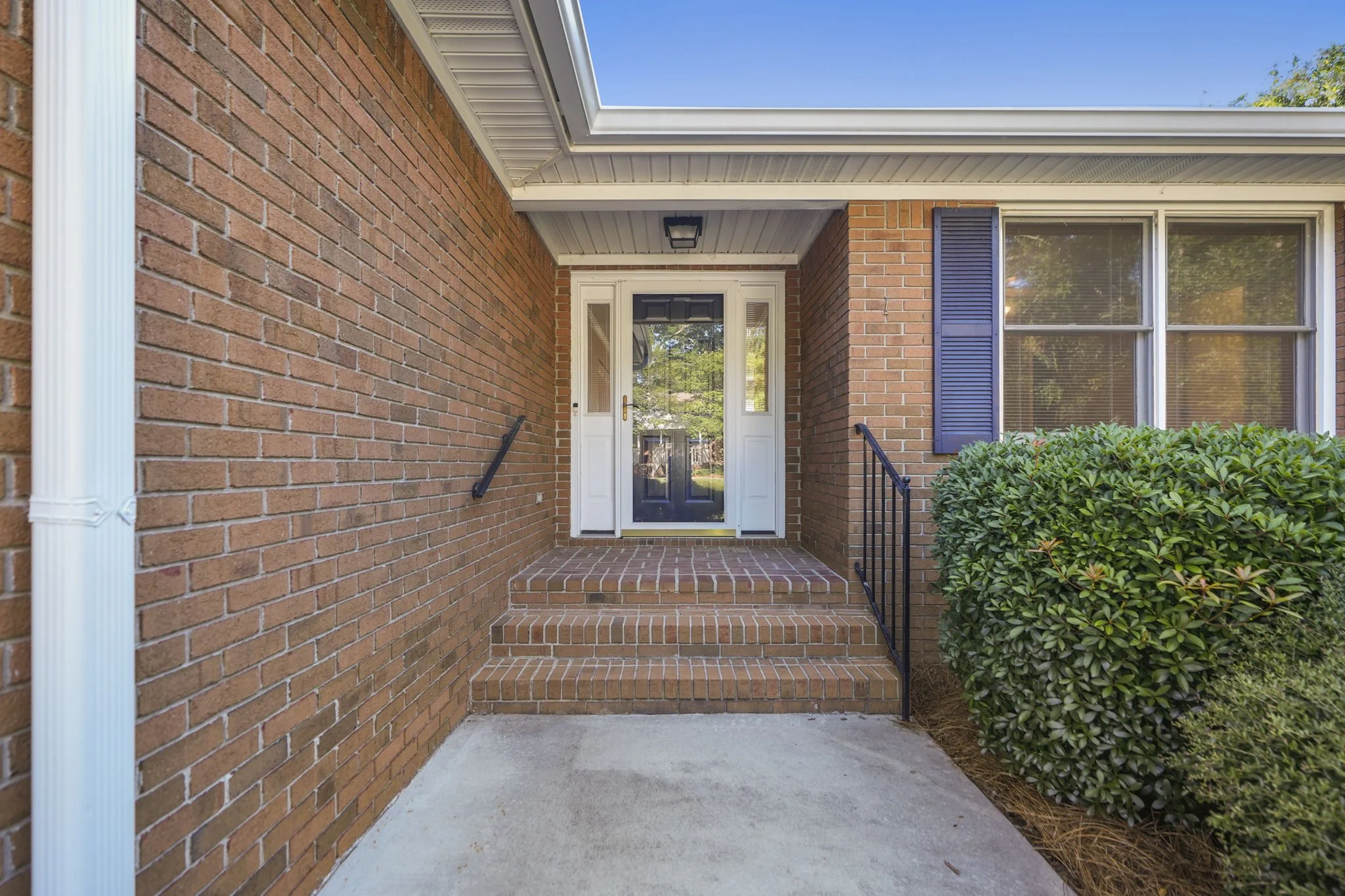 Brick front porch with steps leading to a white front door with sidelights, black railing, a shrub on the right, and a window with blue shutters on the right.