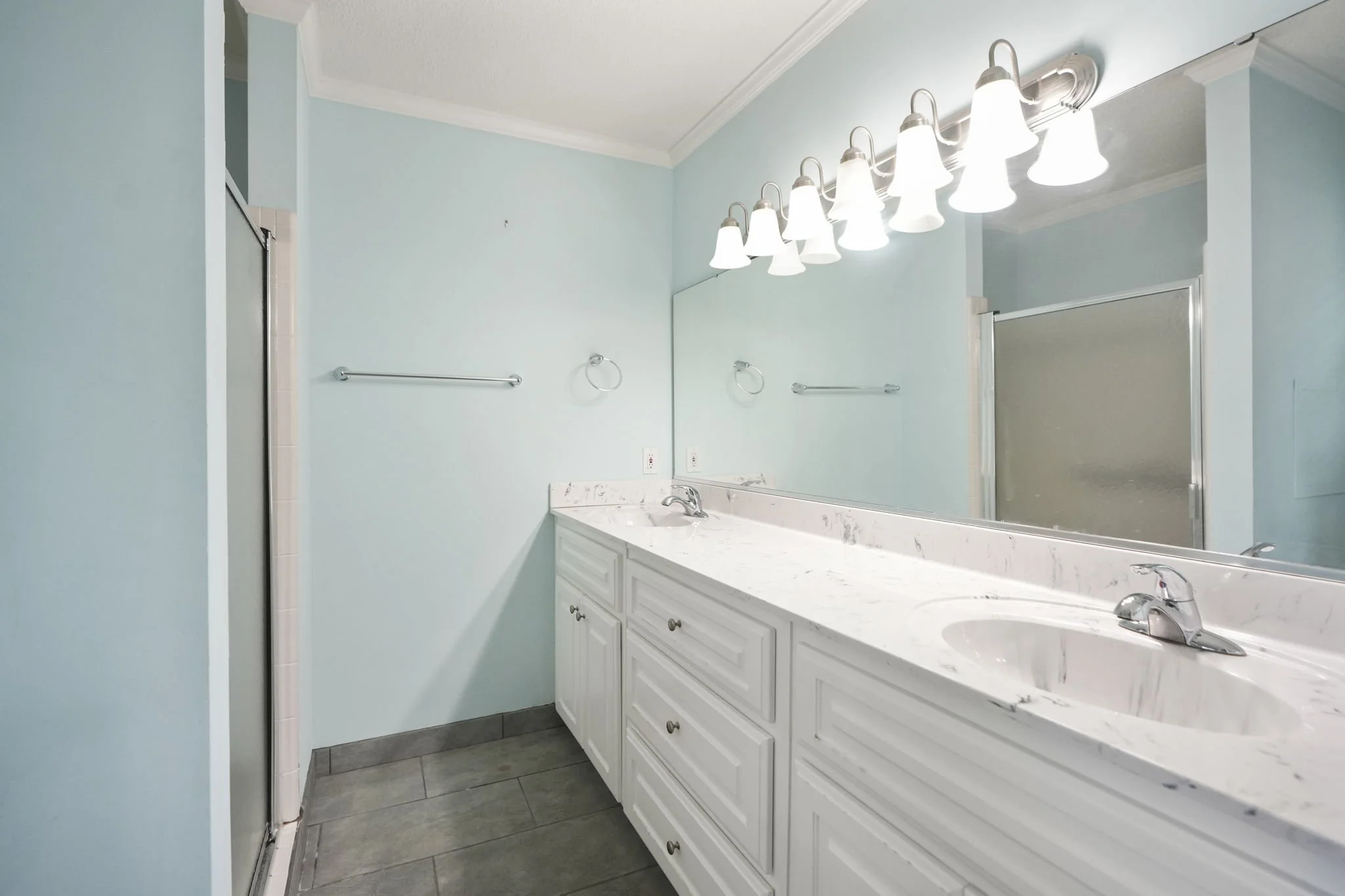 Bathroom with light blue walls, large mirror, white vanity with two sinks, marble countertop, and overhead light fixture with multiple bulbs.