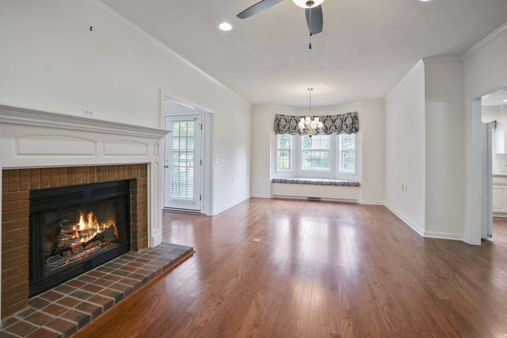 Empty living room with hardwood floors, a brick fireplace, large bay window with curtains and a chandelier, adjacent to a kitchen area.