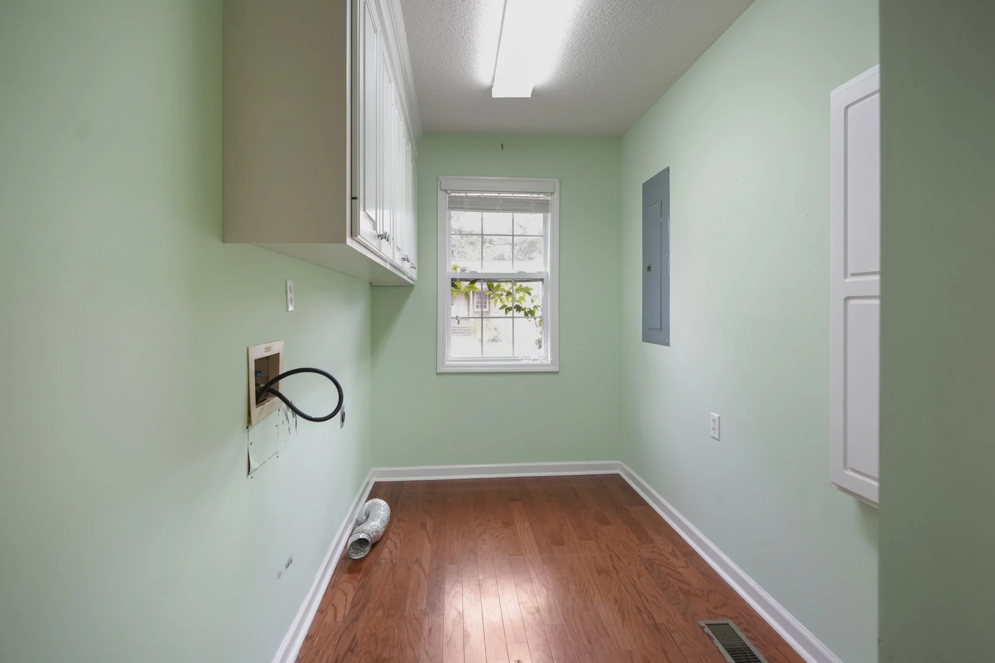 Empty laundry room with light green walls, wooden floor, white cabinets above, a window with blinds, and an electric outlet with a black cord.
