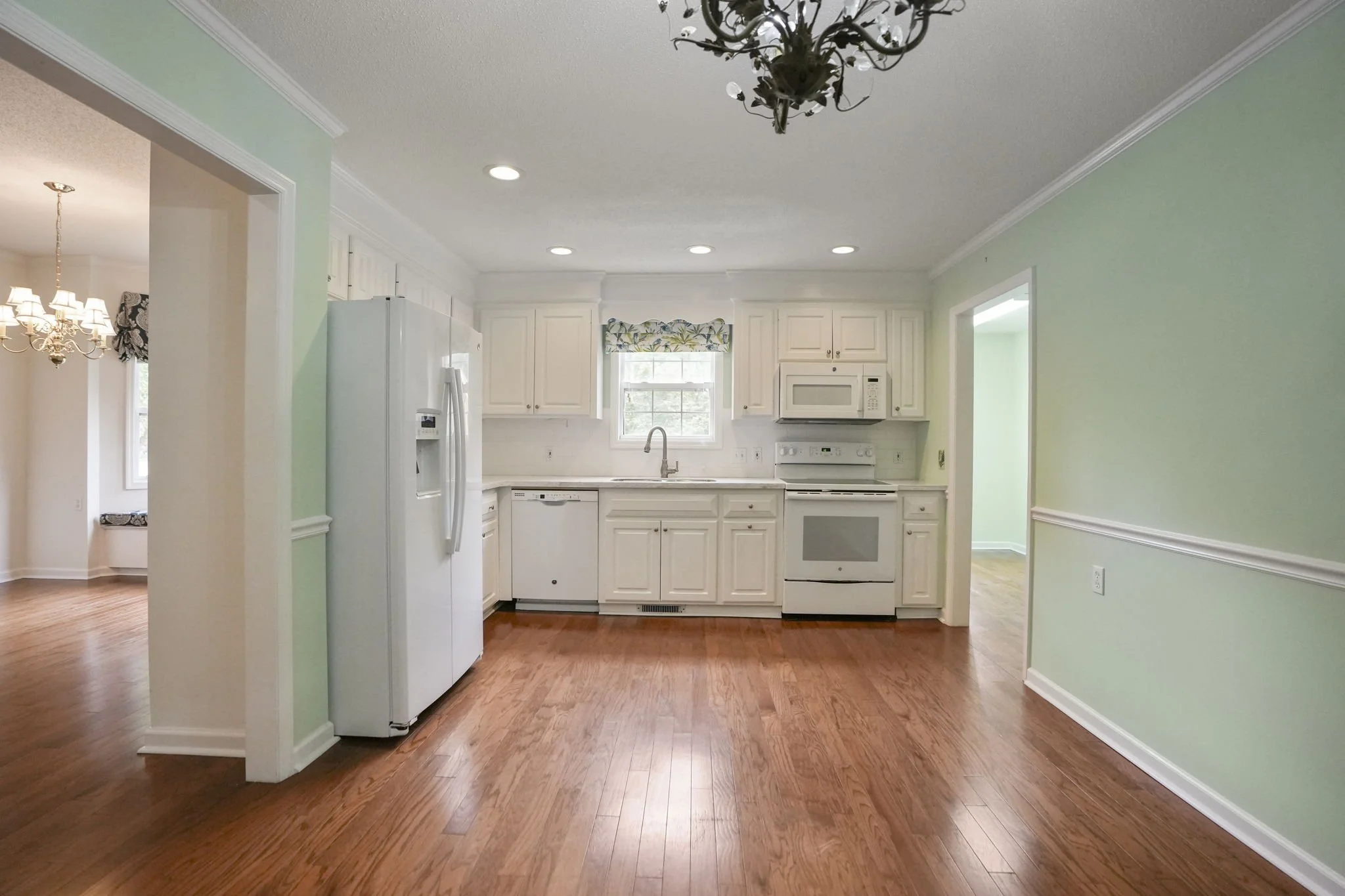 Empty kitchen with white cabinets, white appliances, a small window with a floral valance, hardwood floors, three ceiling lights, and a ceiling-mounted chandelier.