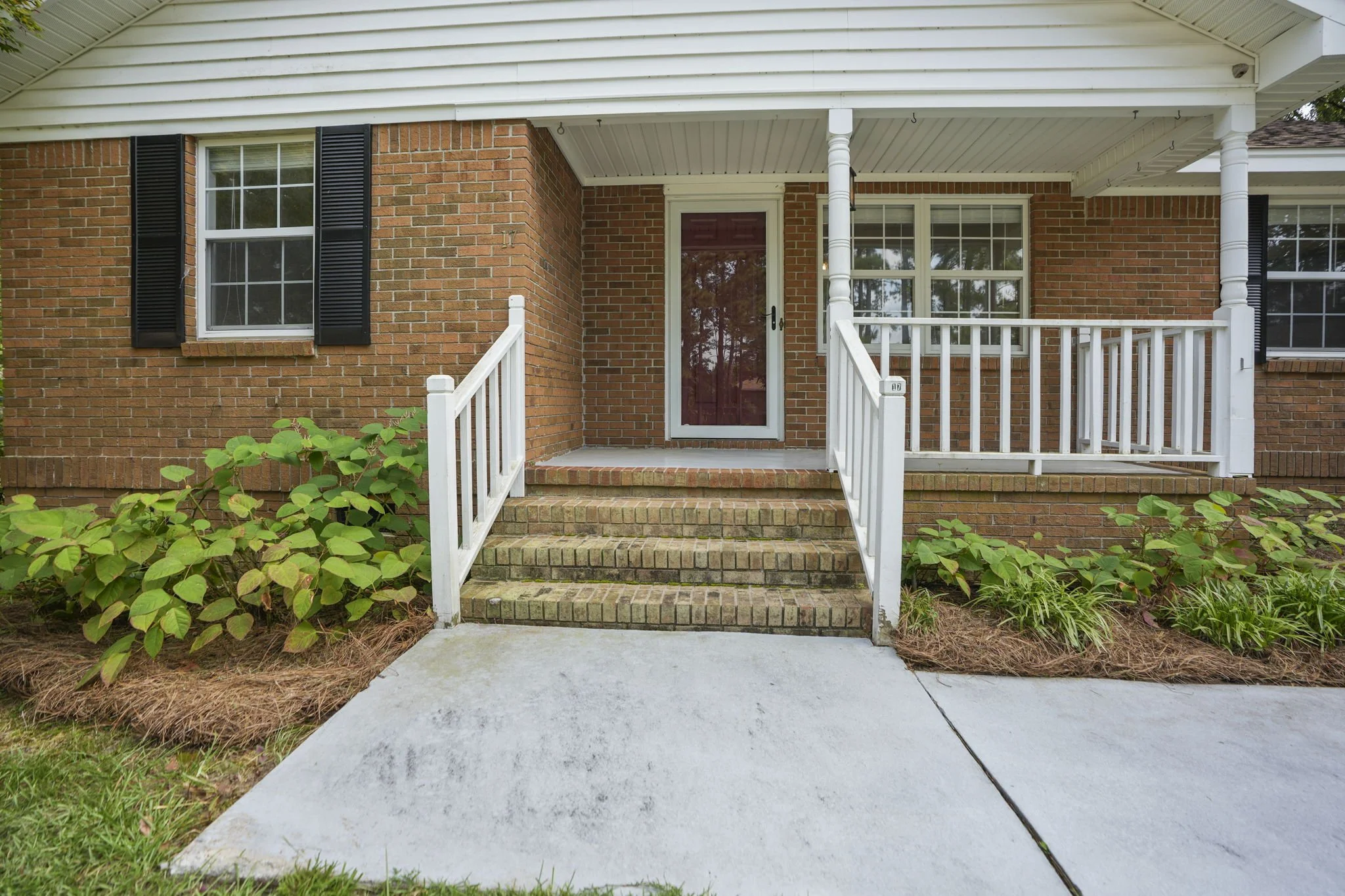 Front porch with brick steps, white railing, and a glass door, outside of a brick house with black window shutters.