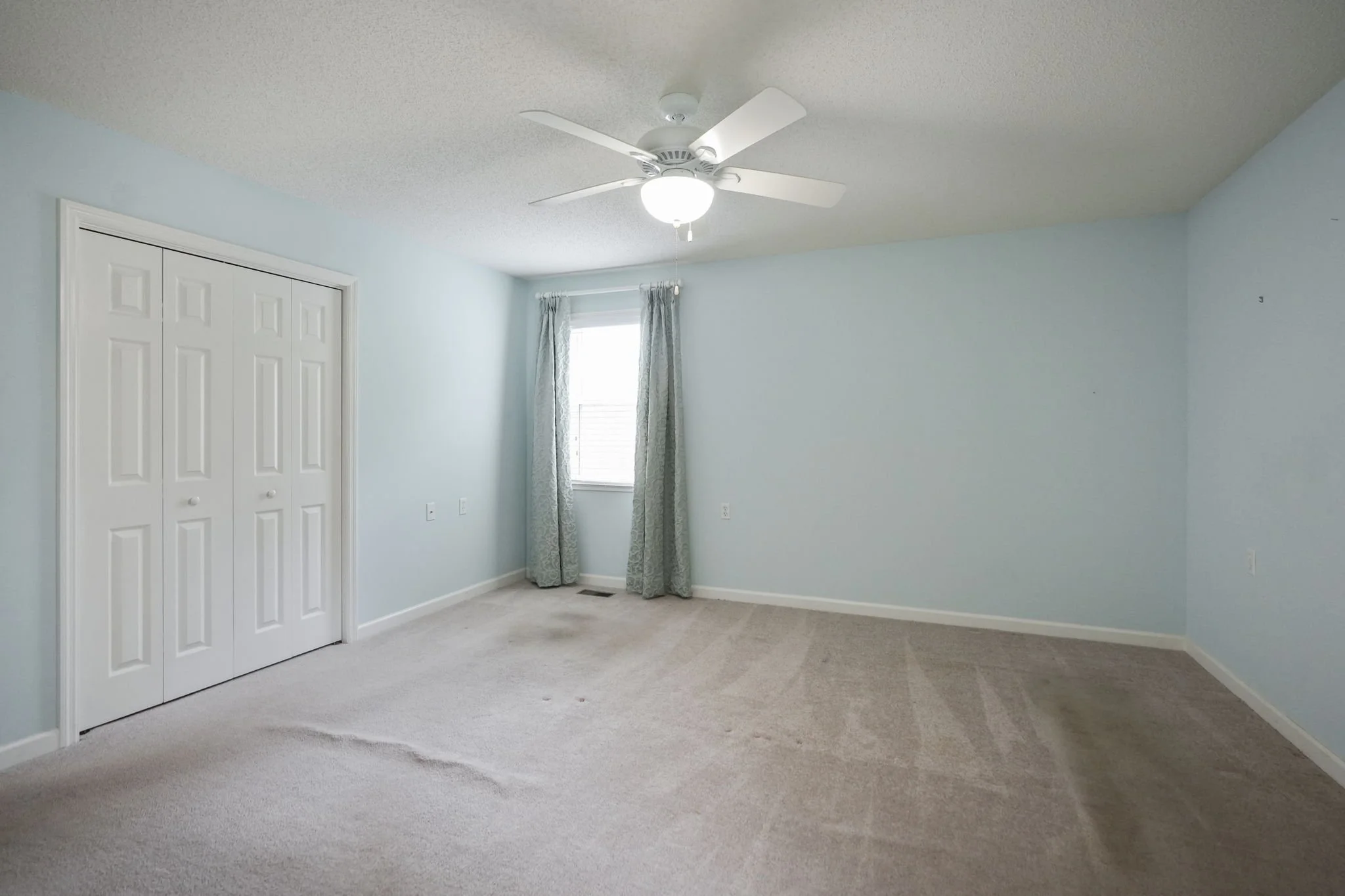 Empty bedroom with light blue walls, beige carpet, a window with patterned curtains, a ceiling fan with light, and a closet with white doors.