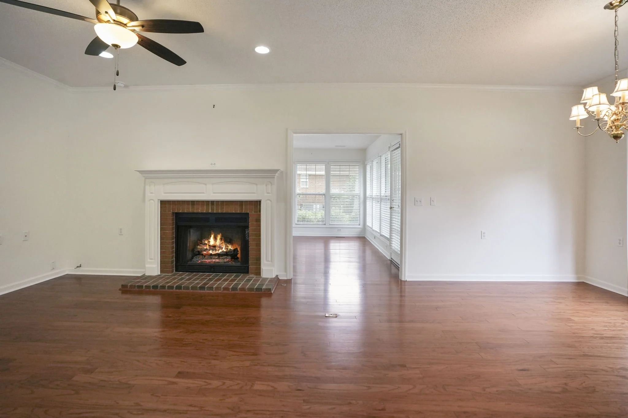 Living room with a lit fireplace, hardwood floors, ceiling fan, and a separate sunroom area with large windows and blinds.