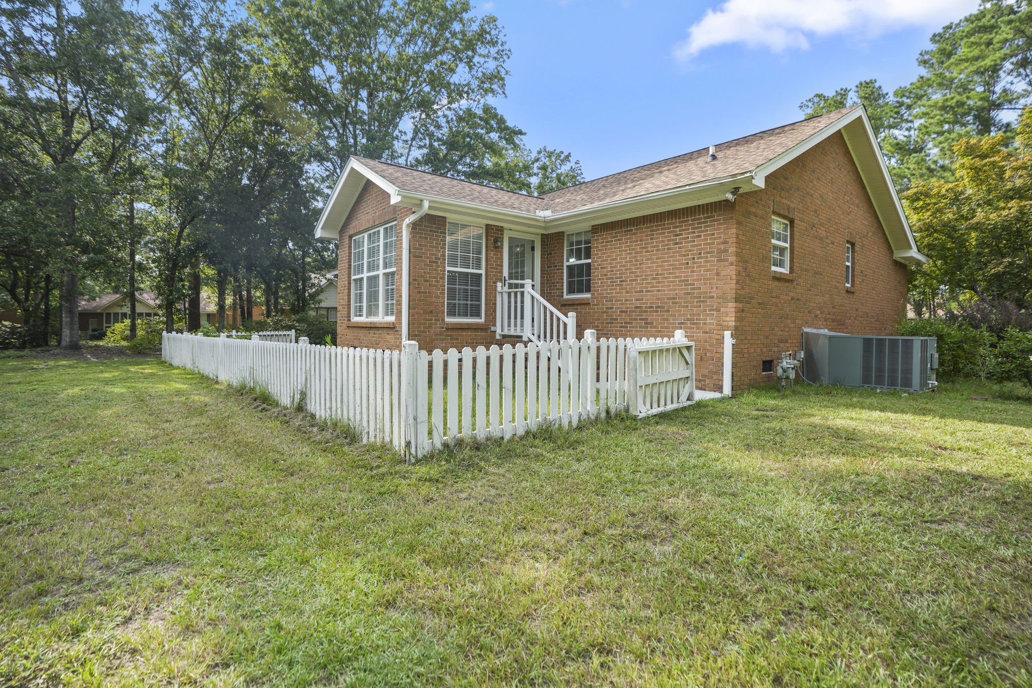 Backyard view of a brick house with white picket fence, surrounded by green grass and trees, under a blue sky.