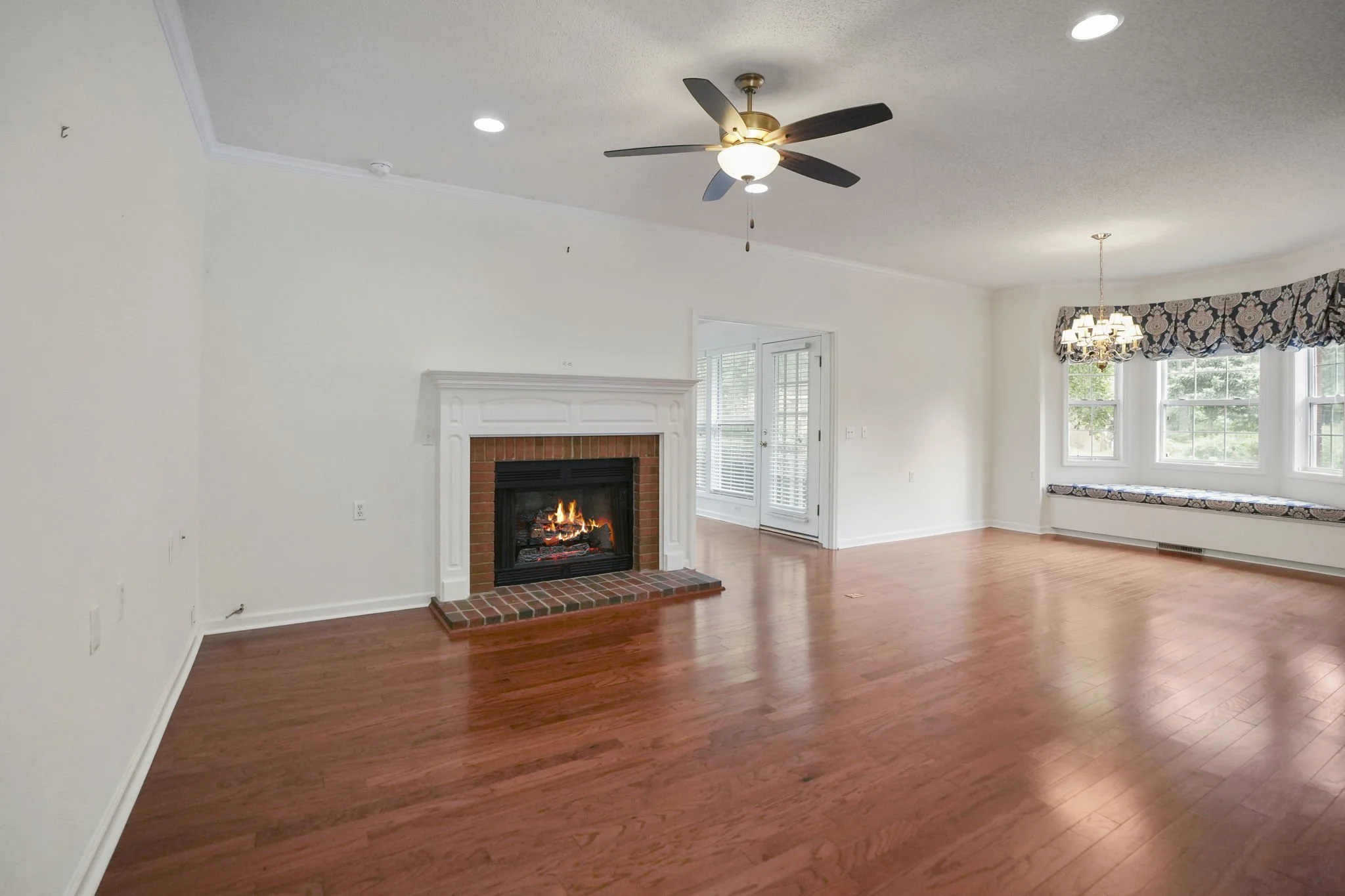 Empty living room with hardwood floors, a fireplace with a brick hearth, a ceiling fan, and large bay windows with patterned valance.