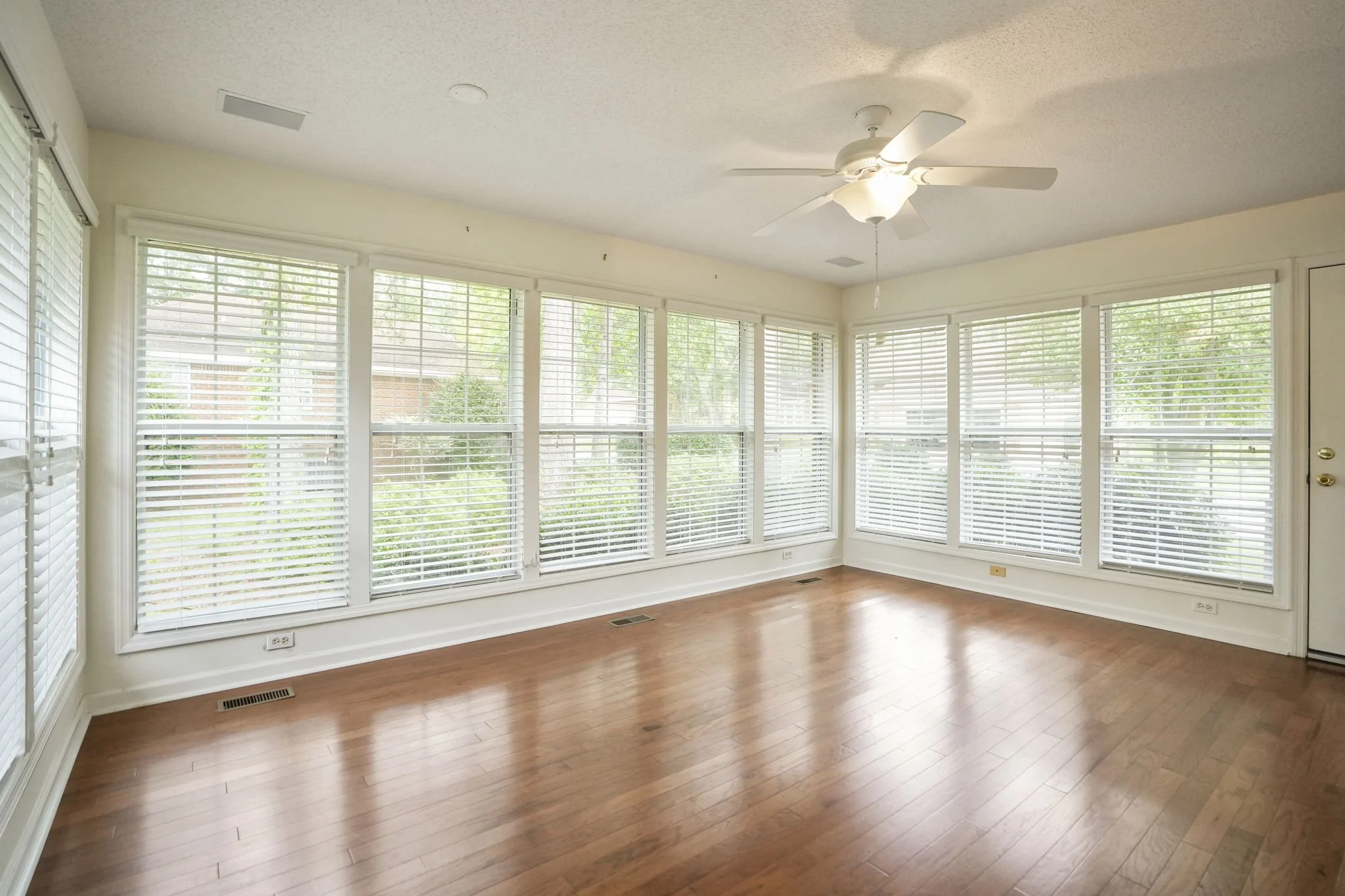 Empty room with large windows, hardwood floors, white walls, a ceiling fan, and blinds on the windows.