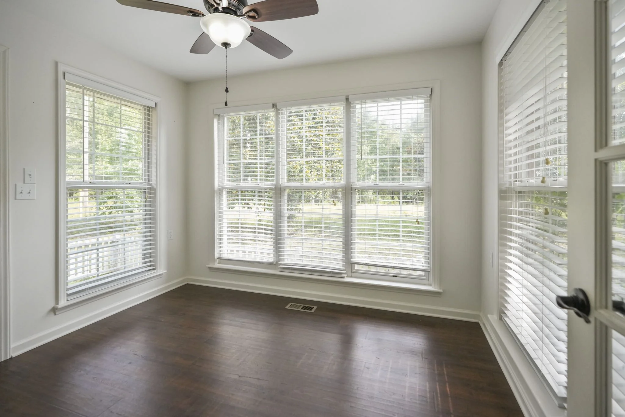 Empty room with large windows, white walls, hardwood floor, ceiling fan with light fixture.