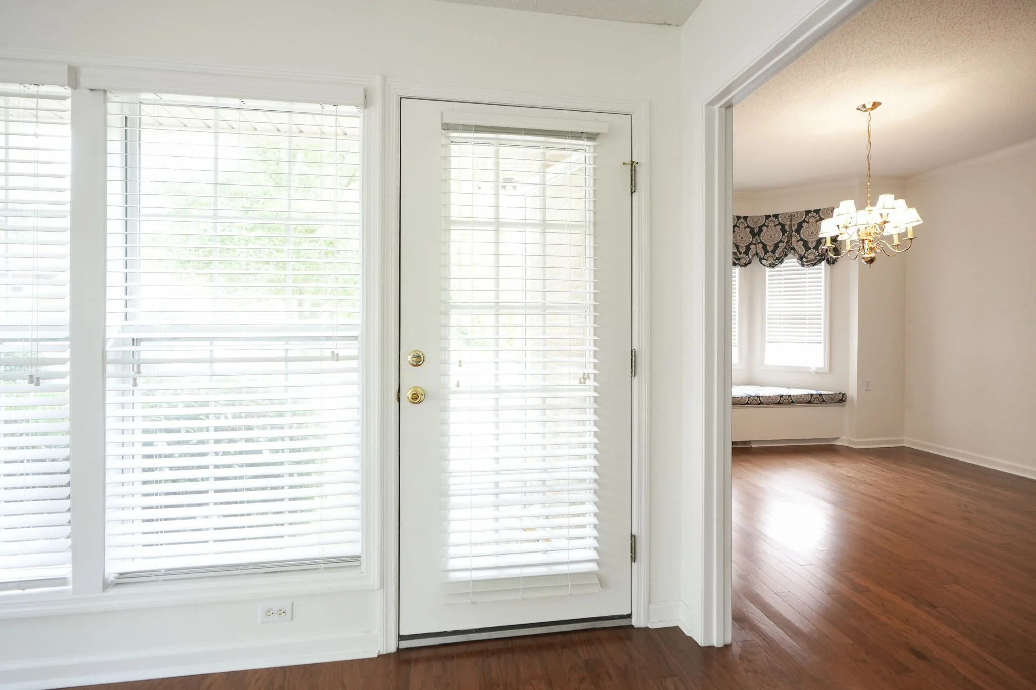 Interior view of a bright room with a door with blinds leading outside, and a view into another room with a chandelier and a window with a valance, and hardwood floors.