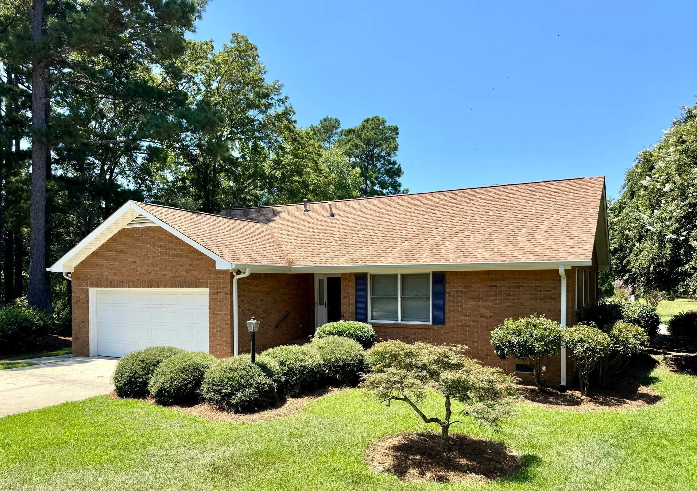 A single-story brick house with a brown shingle roof, white garage door, and front yard with shrubs and a small tree. Clear blue sky and tall trees in the background.