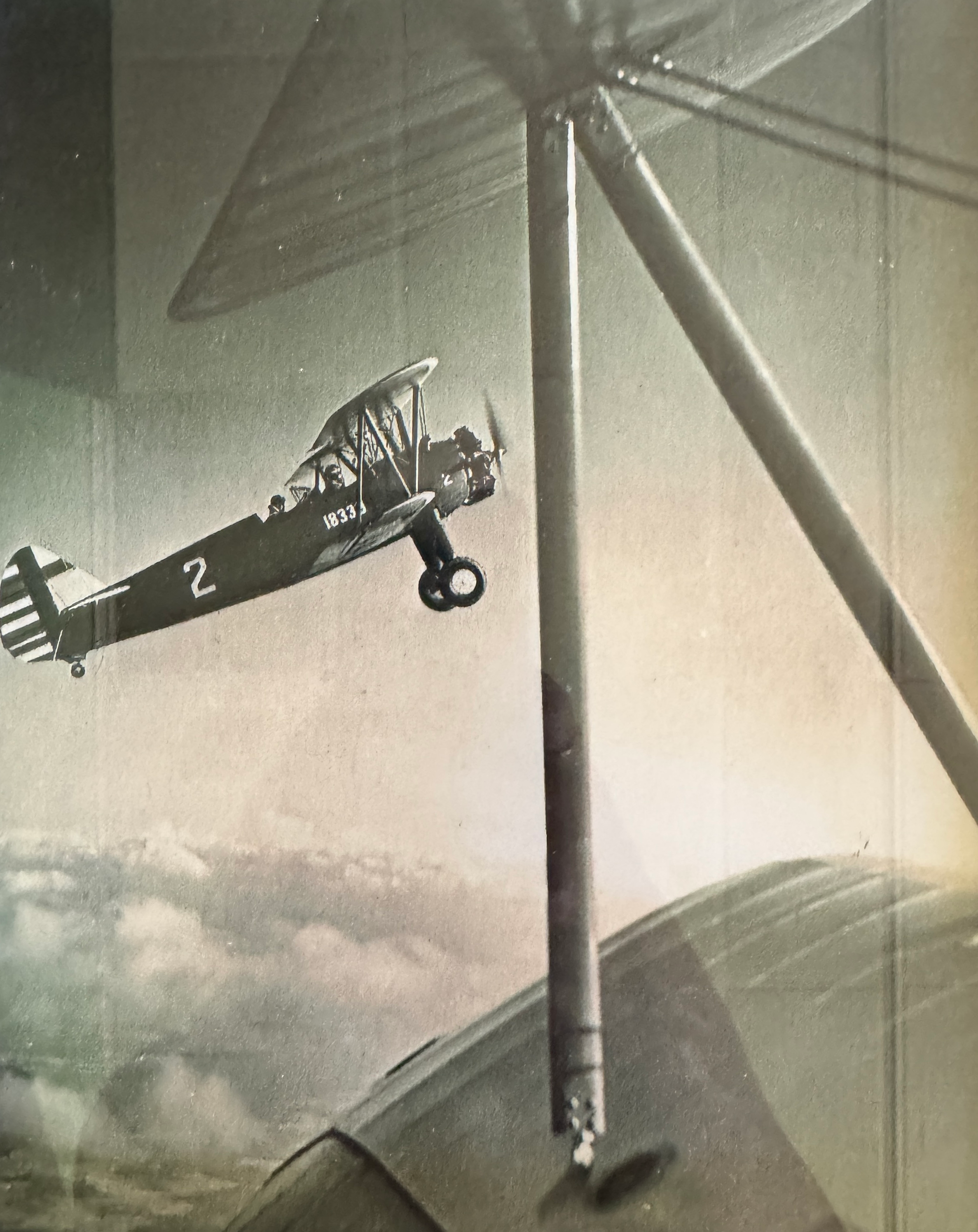 A vintage black and white photograph of a biplane aircraft flying in the sky, with the nose pointing slightly upward, and the number 2 visible on the side of the fuselage.