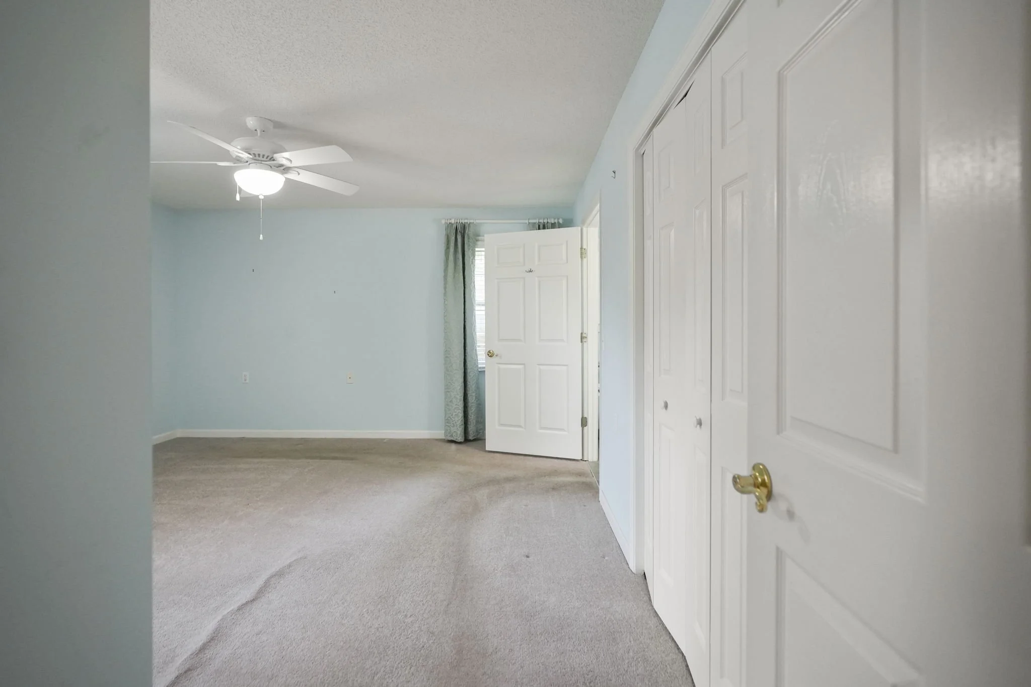 An empty bedroom with light blue walls, beige carpet, ceiling fan, white closet doors, open door with curtains, and a window with blinds.