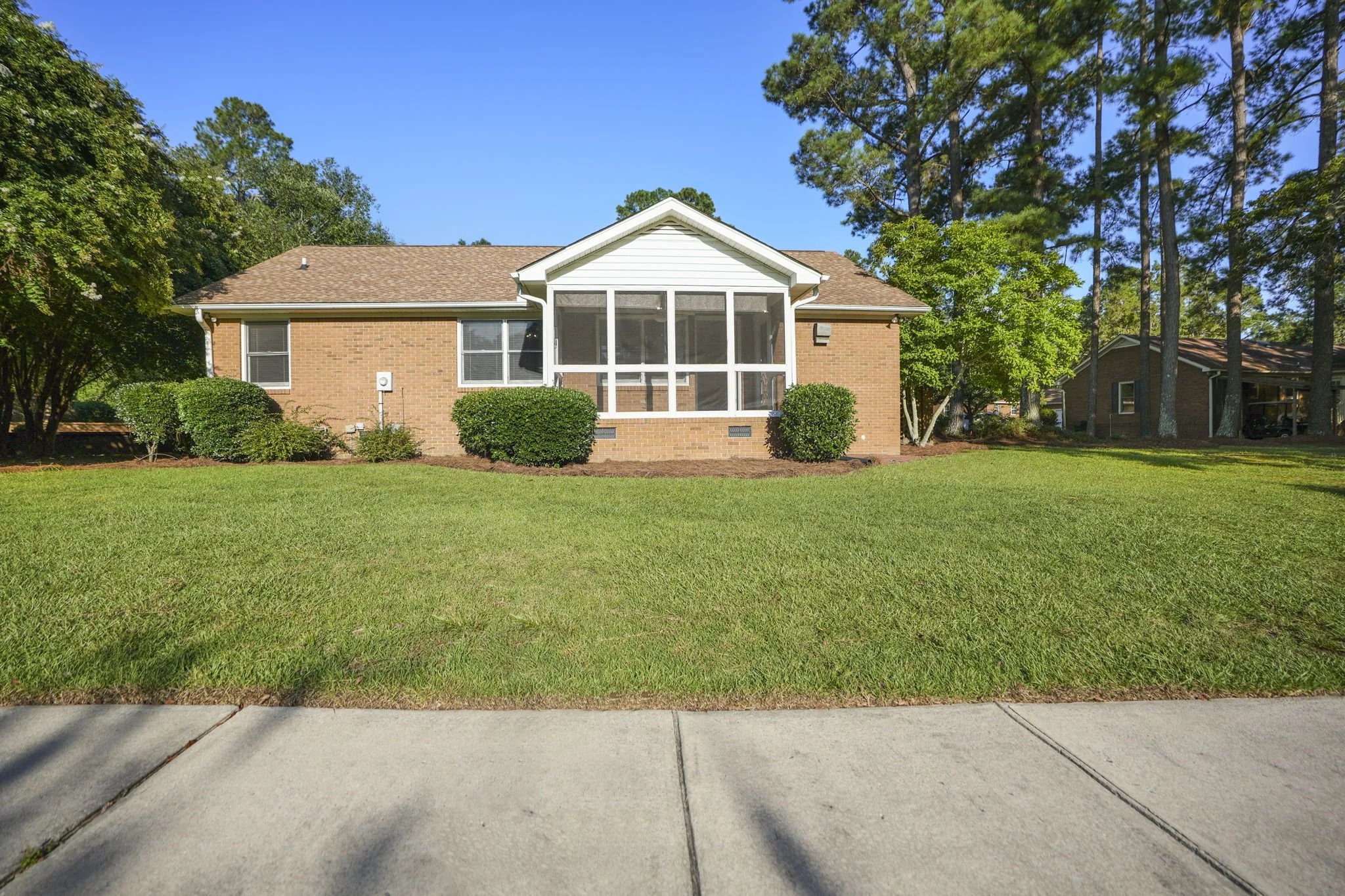 A brick house with a screened porch, green lawn, and trees under a clear blue sky.
