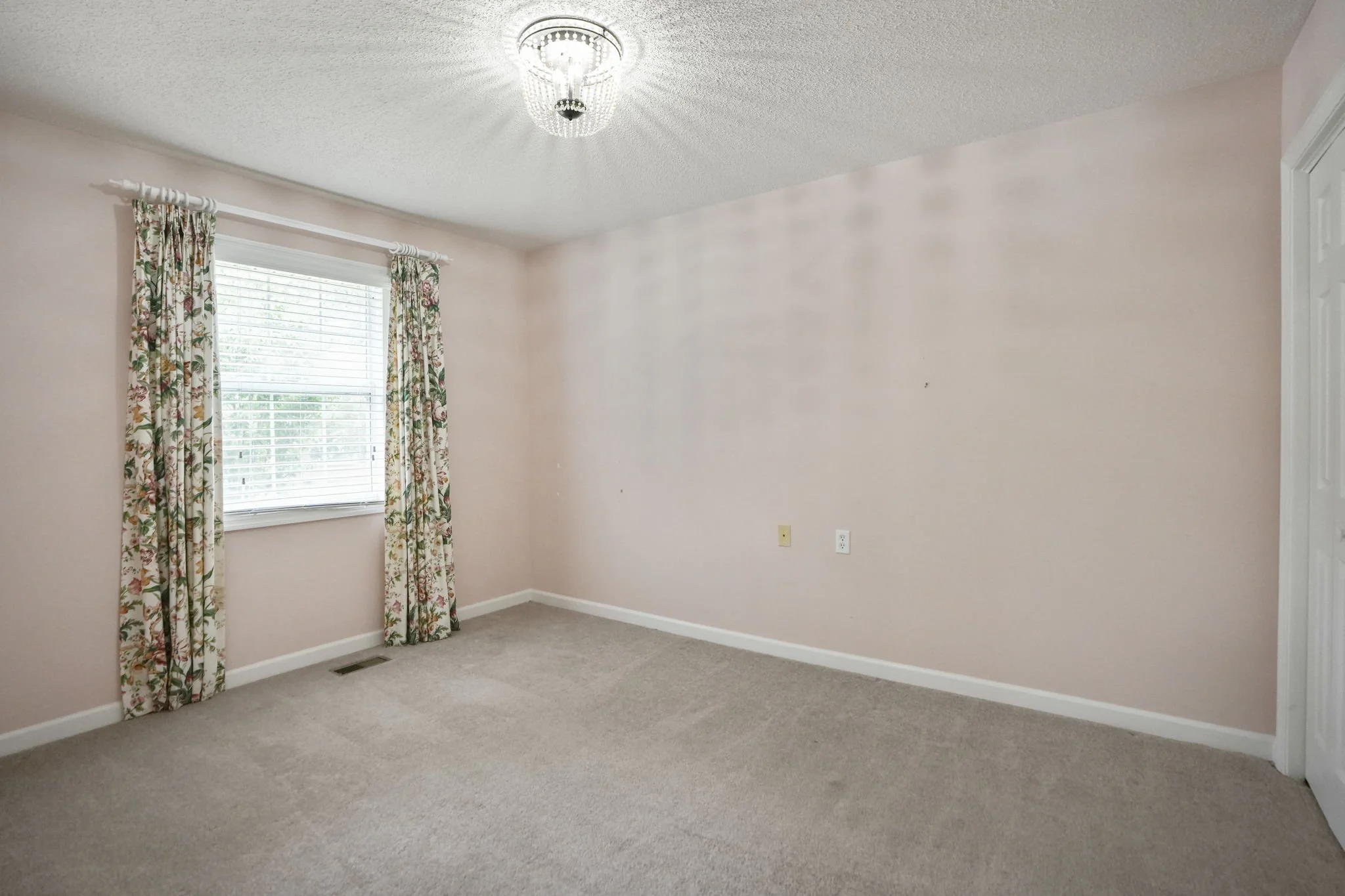 Empty room with pink walls, window with floral curtains, beige carpet, ceiling light fixture, and a partially open door.