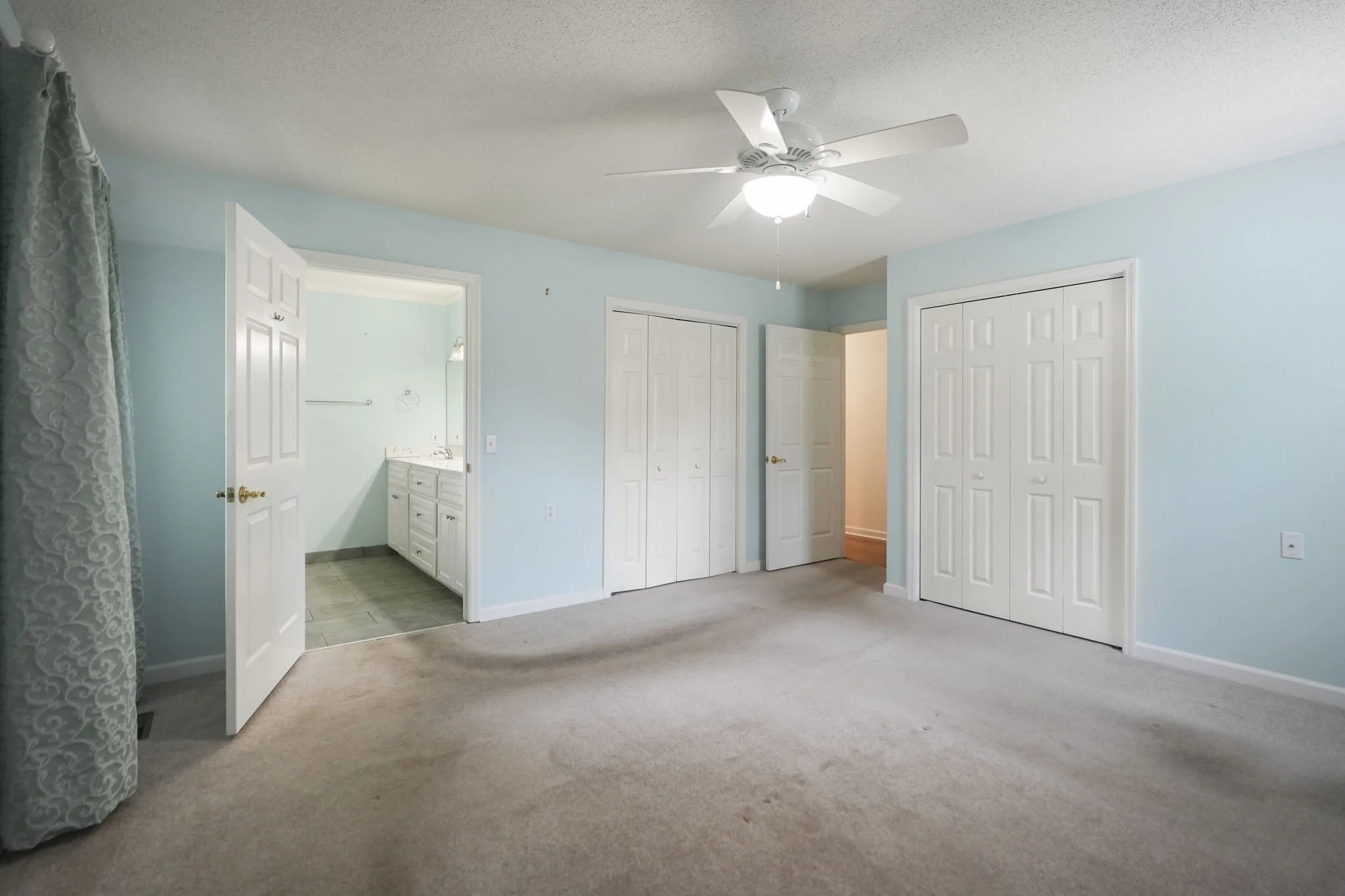 Empty bedroom with light blue walls, beige carpet, white closet doors, ceiling fan with light, open door leading to bathroom, and a window with patterned curtains.