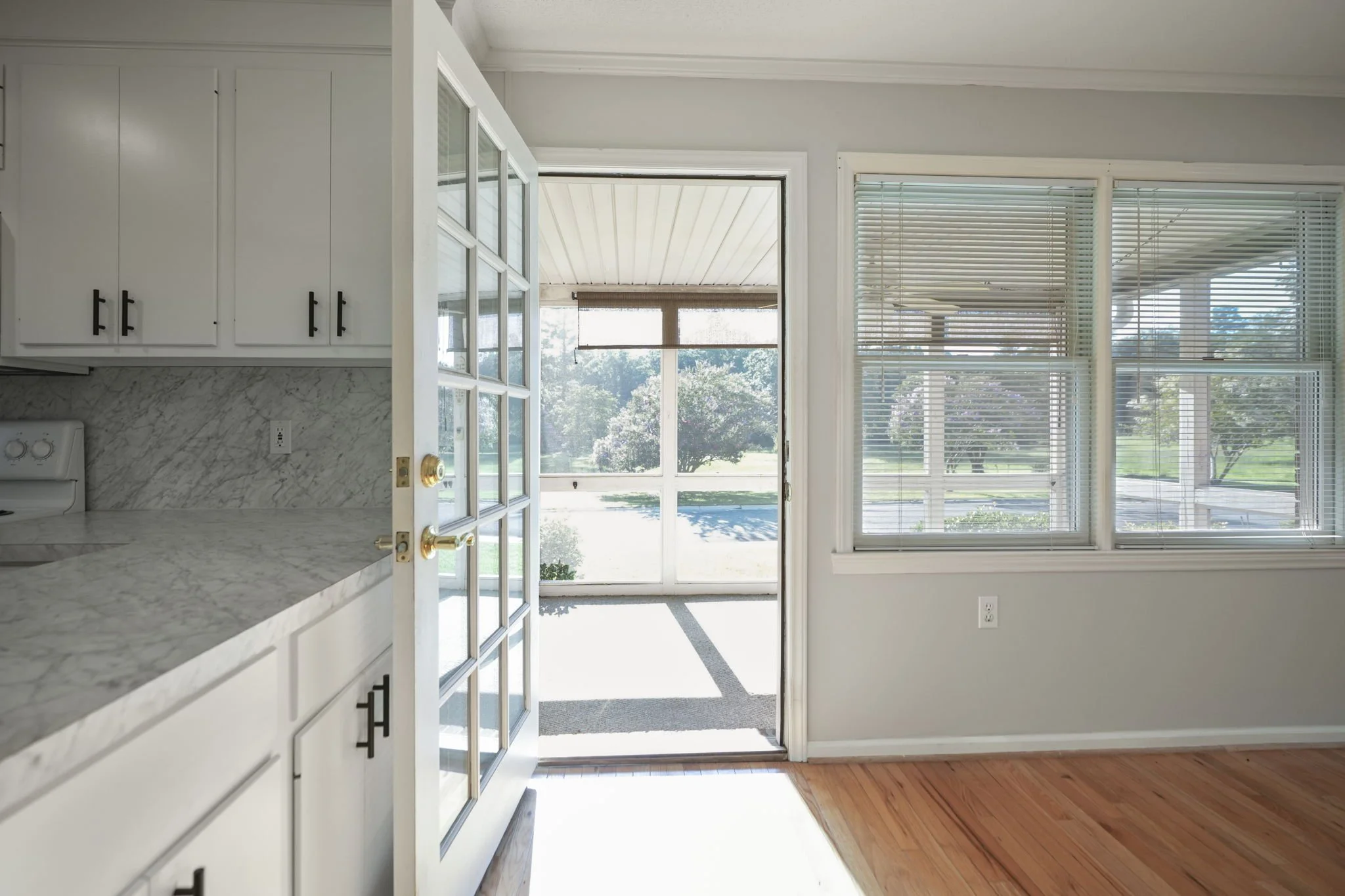 Interior view of a kitchen with white cabinets, marble countertops, and a glass door leading to an enclosed porch with large windows showing outdoor greenery.