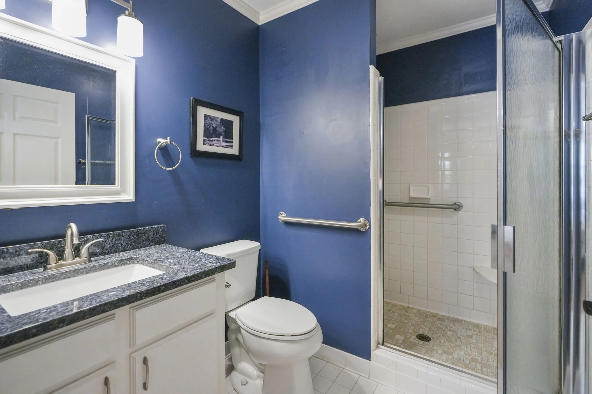 Bathroom with blue walls, a white vanity with a granite countertop, a mirror, a picture hanging, a toilet, and a walk-in shower with white tiles and a grab bar.