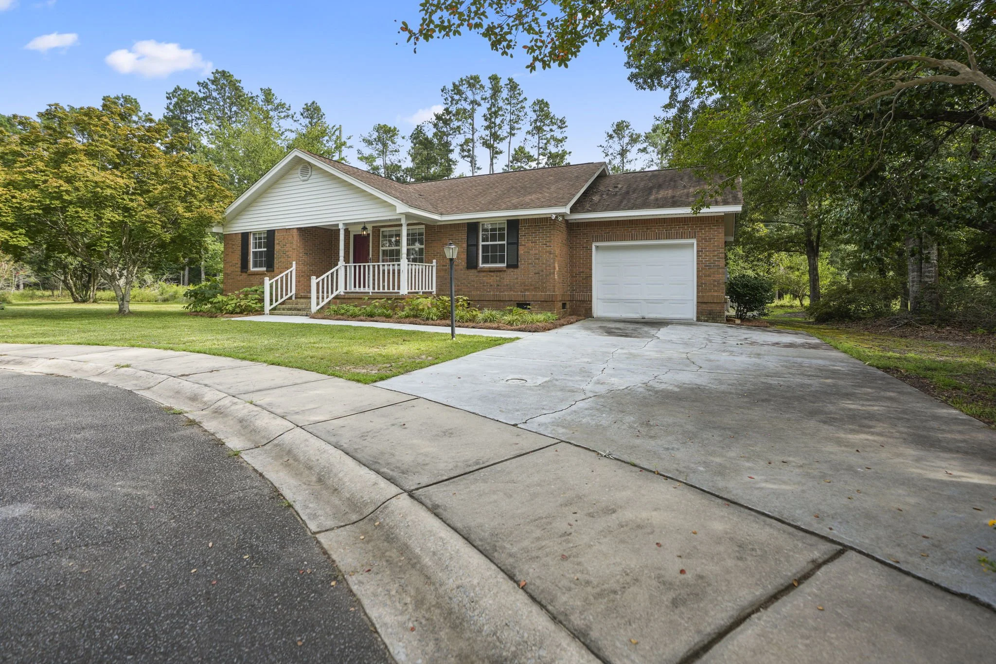 Front view of a single-story brick house with a small porch, white railing, and a driveway leading to an attached garage. The house is surrounded by a grassy yard and trees.