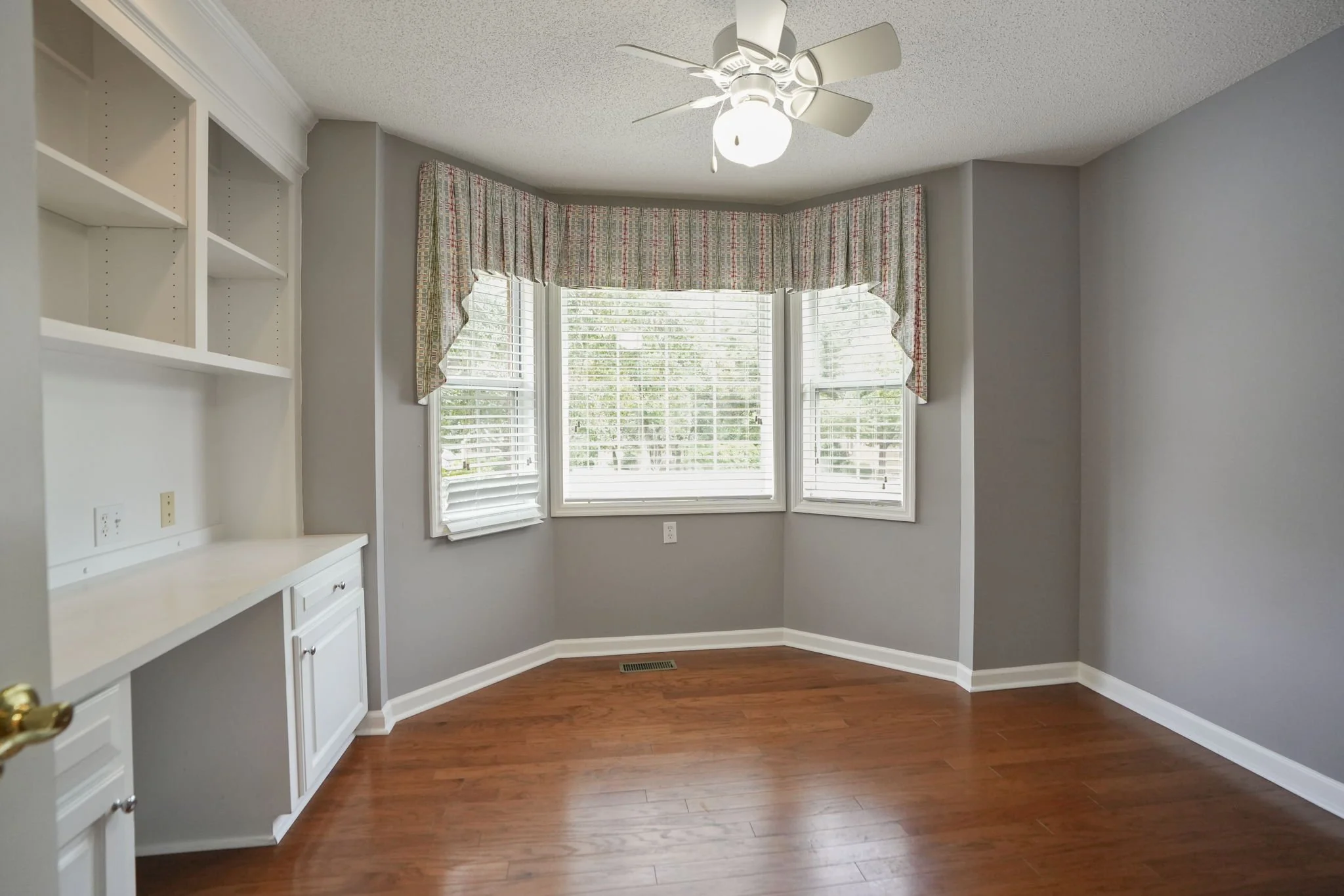 Empty room with hardwood floor, gray painted walls, a corner window with a valance, ceiling fan with light, and built-in white shelves.
