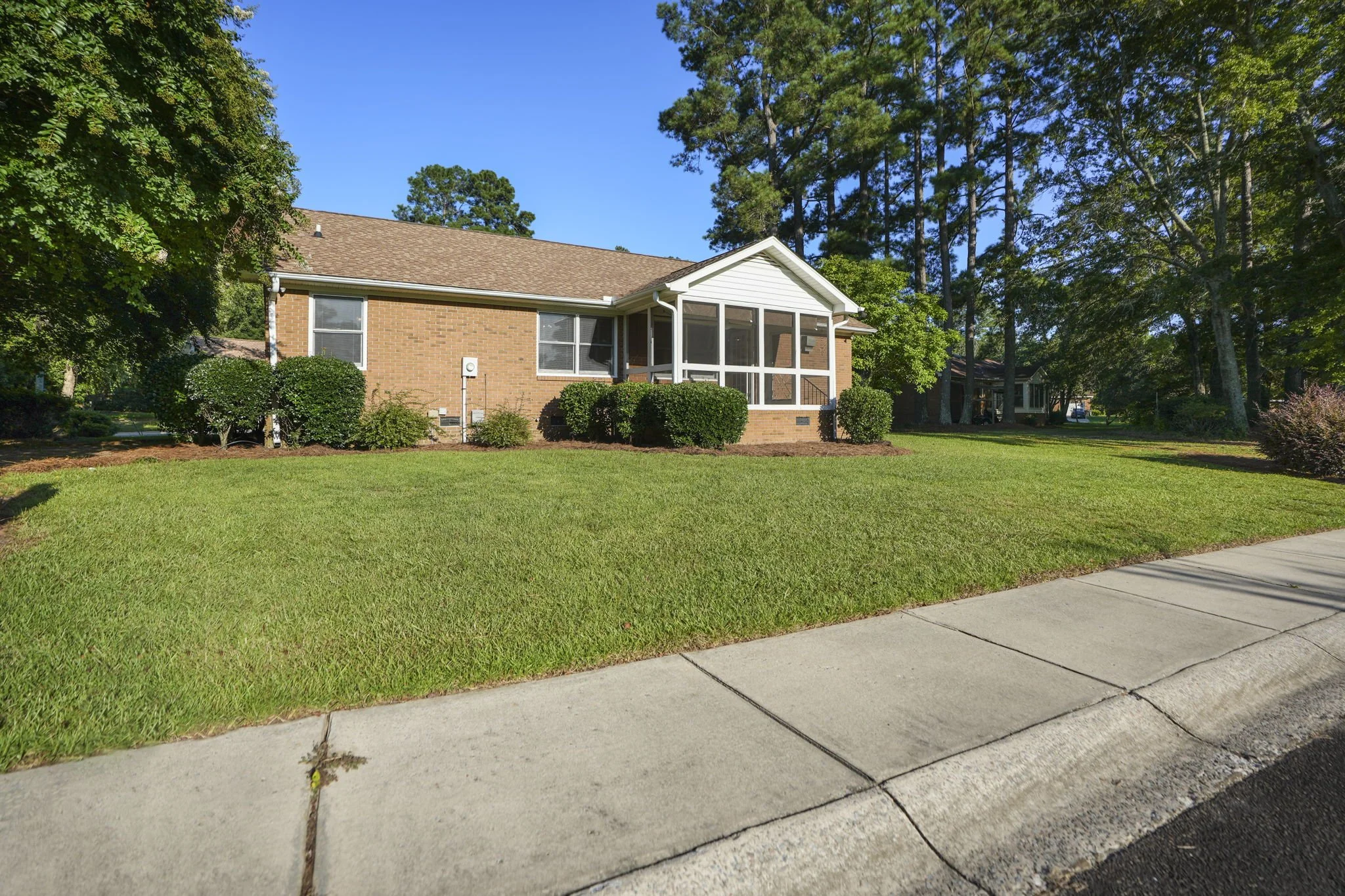 Front view of a brick house with a screened porch, surrounded by manicured lawn and bushes, with tall trees in the background and a concrete sidewalk in the foreground.