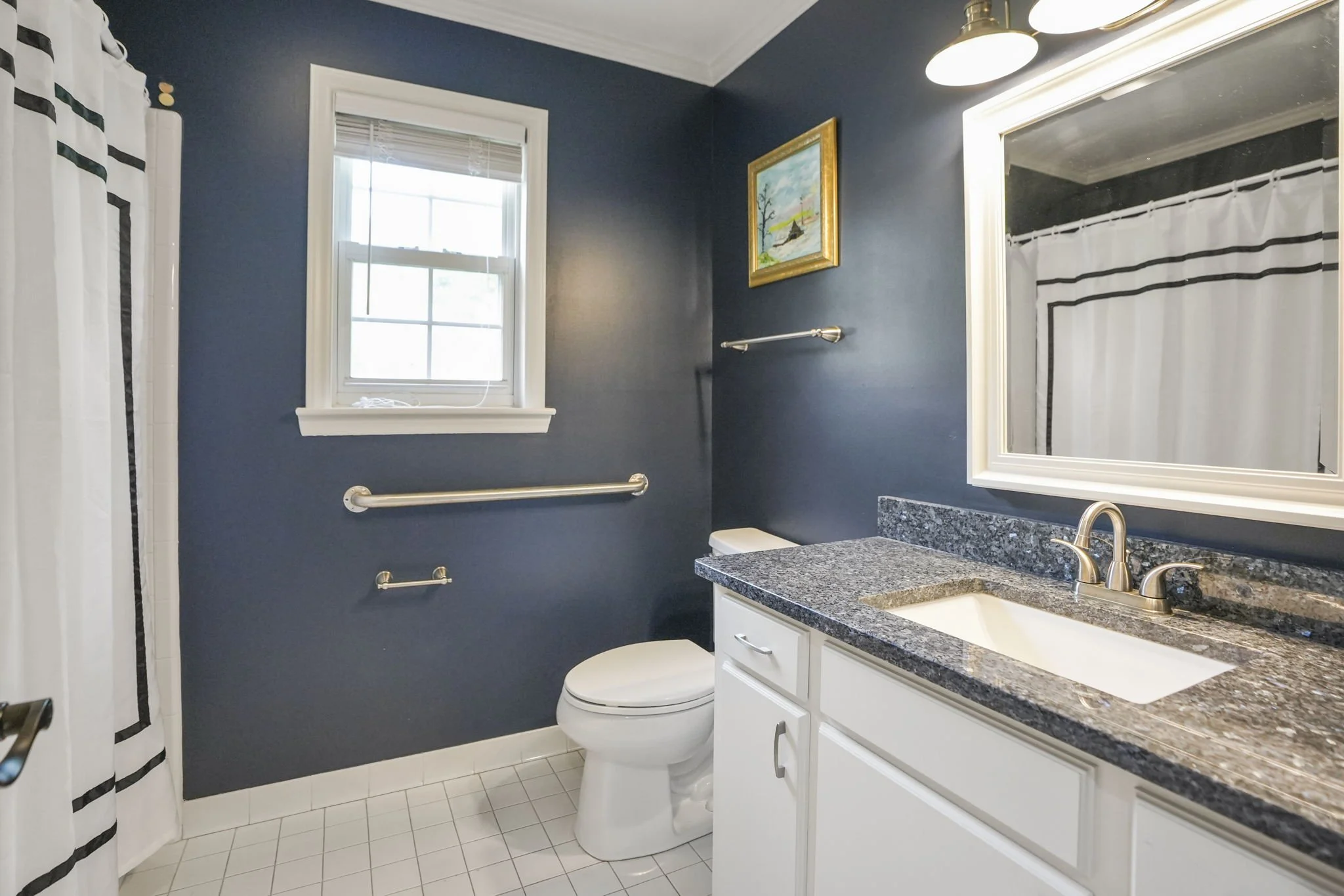 Bathroom with navy blue walls, white tile floor, white vanity with granite countertop, rectangular mirror, small window with blinds, toilet, and white shower curtain with black trim.