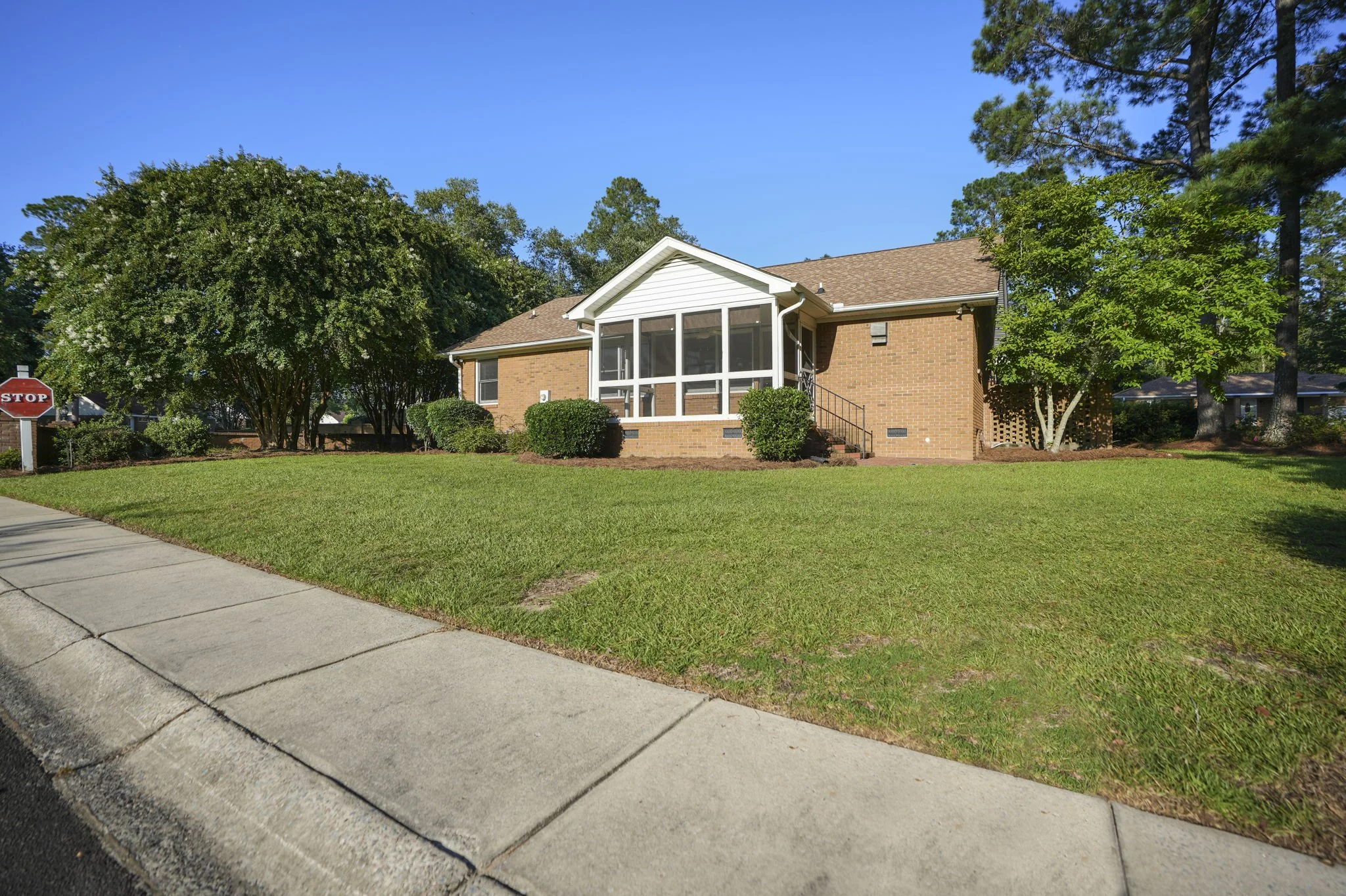Brick house with a screened porch, surrounded by green grass and trees on a sunny day.