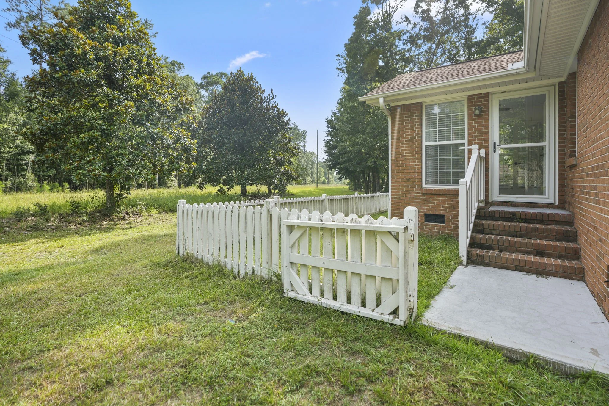 Backyard with a white picket fence, small gate, brick steps leading to a screened porch door, brick house, and large trees in the background under a blue sky.
