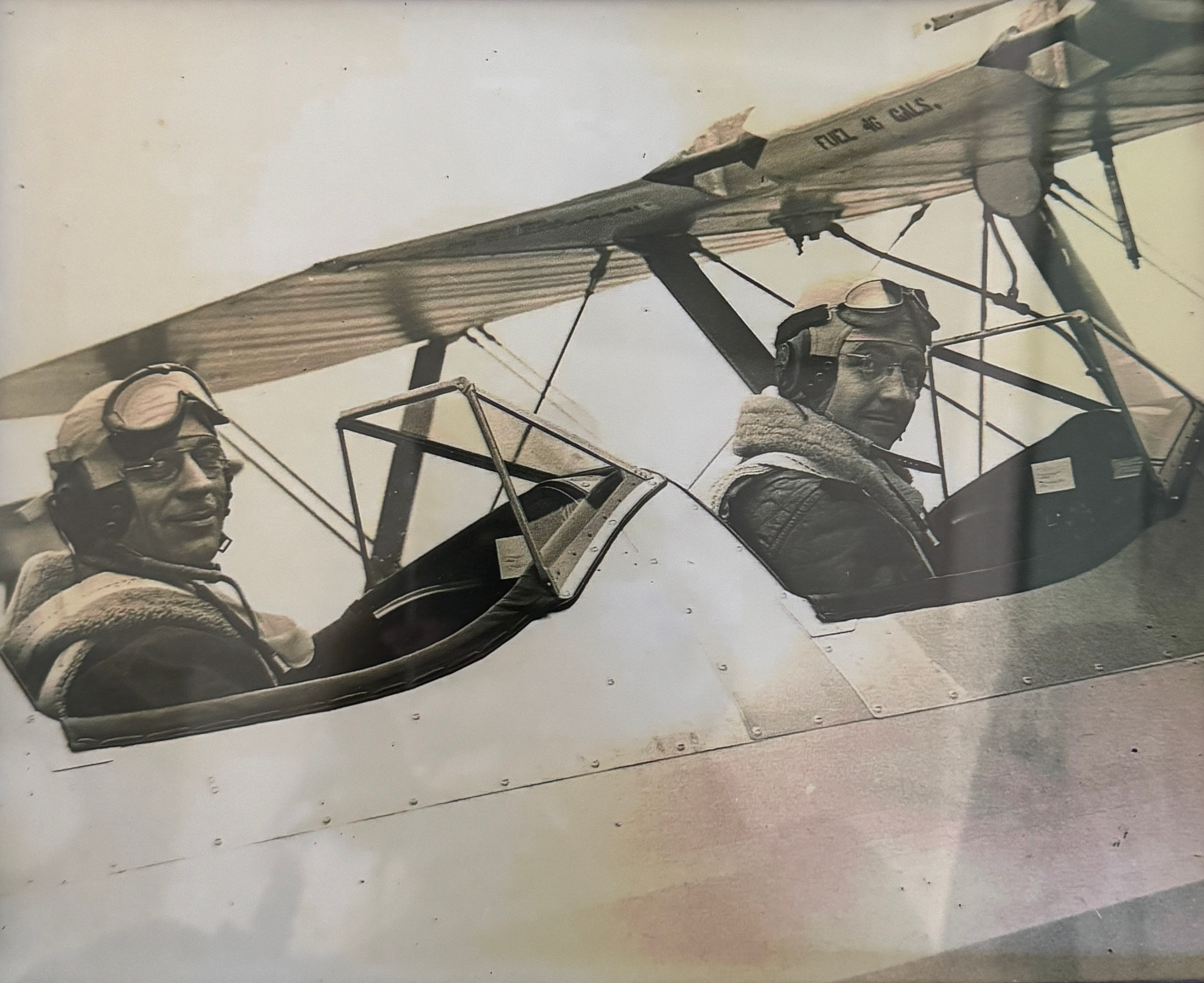 Black and white vintage photograph of two pilots in a biplane. The pilots are wearing leather jackets, goggles, and headsets, seated in the open cockpit, with the plane's wing above them.