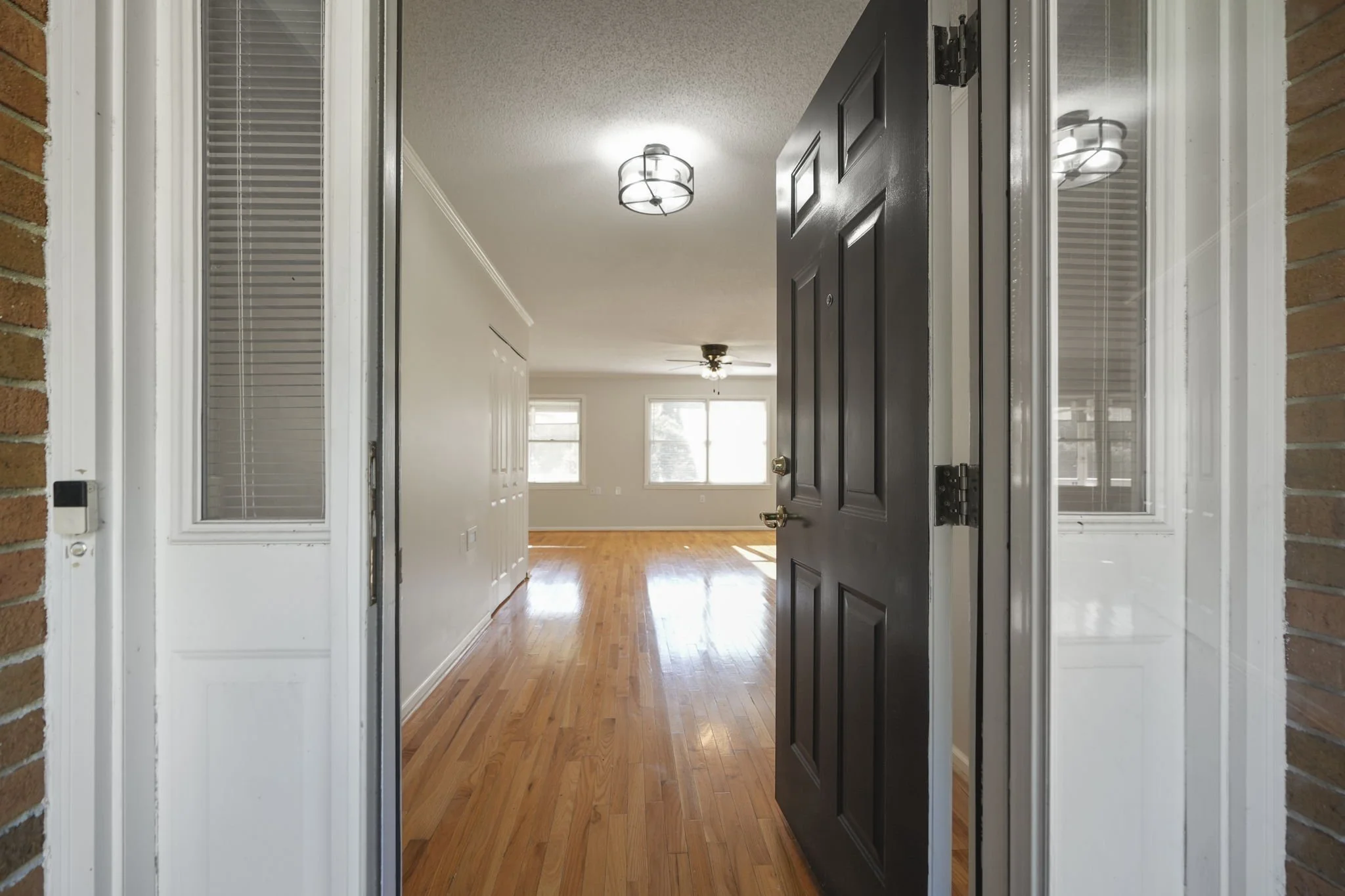 View from front door into an empty living room with hardwood floors, white walls, large windows, ceiling fan, and ceiling light fixtures.