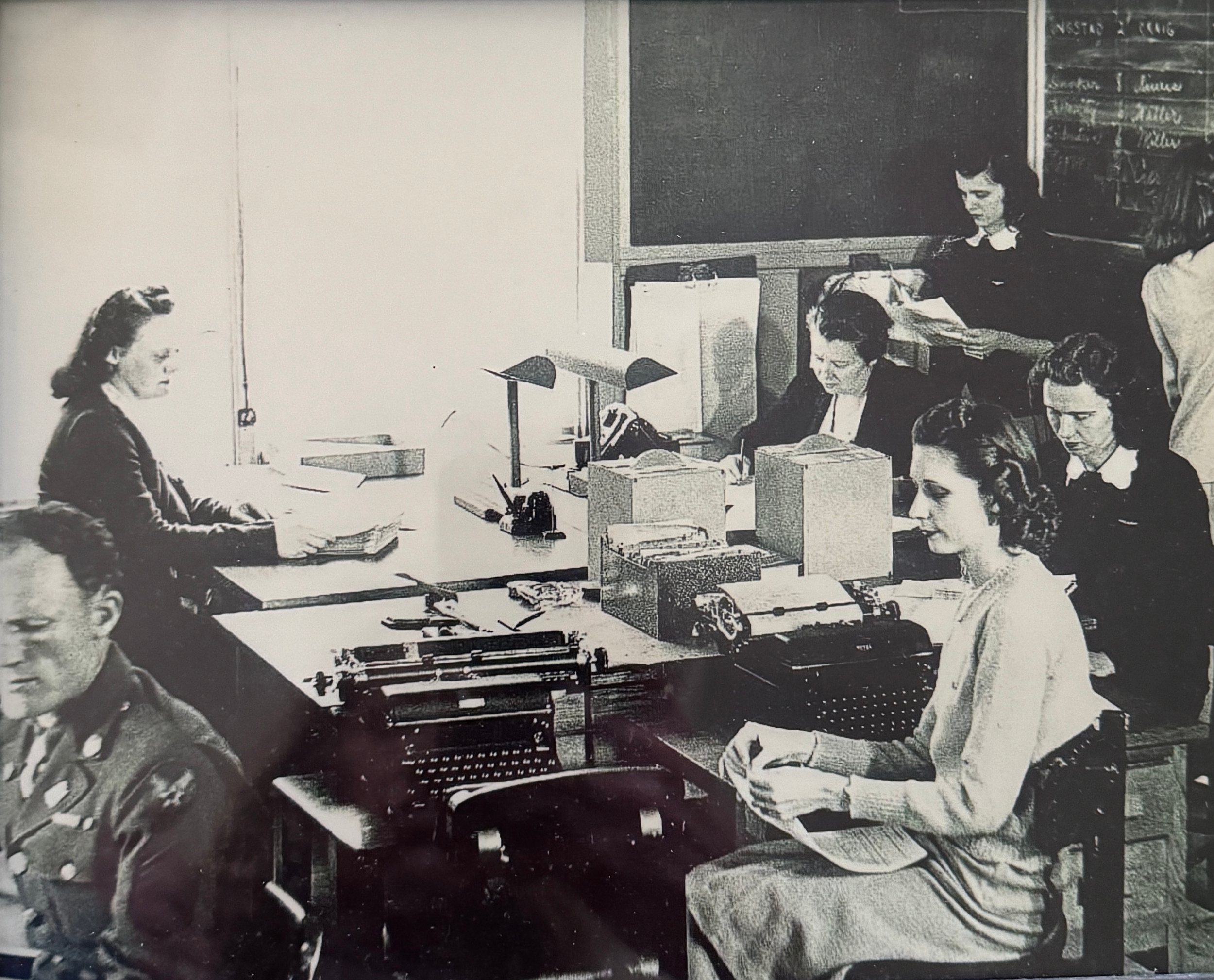 A black-and-white photo of women working at desks in an office, some using typewriters, in a classroom or office setting.
