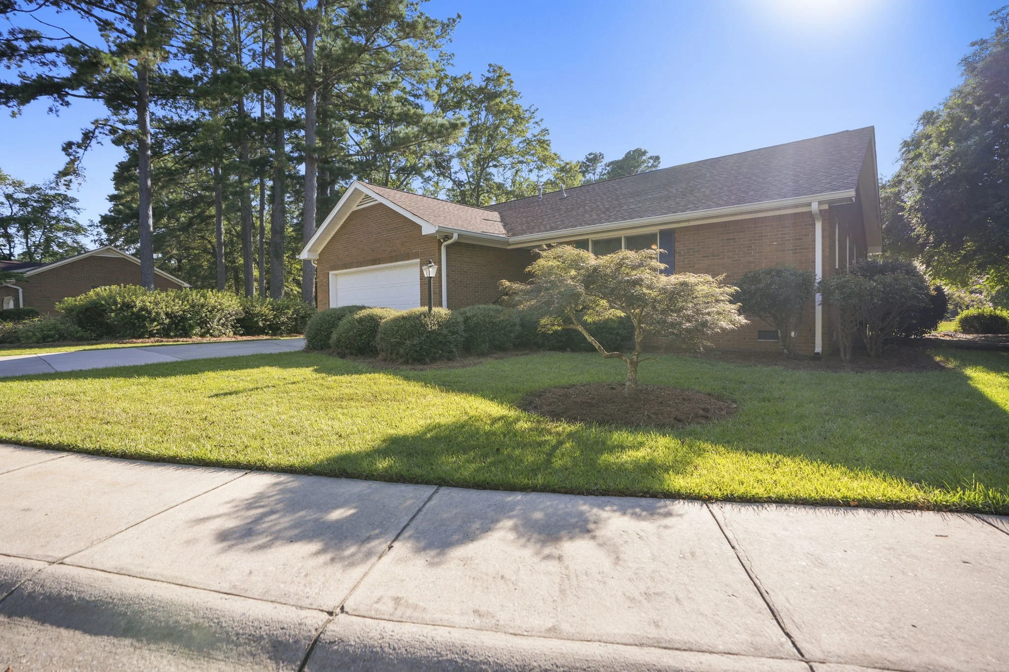 A suburban house with a brick exterior, white garage door, and well-kept front yard with green grass, bushes, and a small tree, under a sunny blue sky.