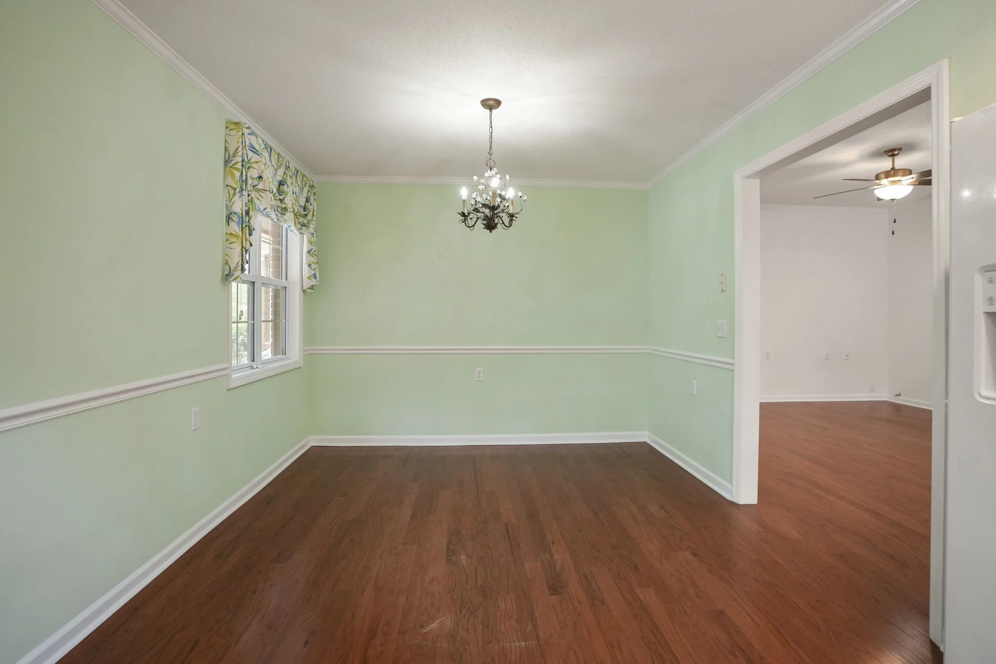 Empty dining room with light green walls, a chandelier hanging from the ceiling, a window with patterned valance curtains, and hardwood floors. An open doorway leads to another room with similar flooring and a ceiling fan.