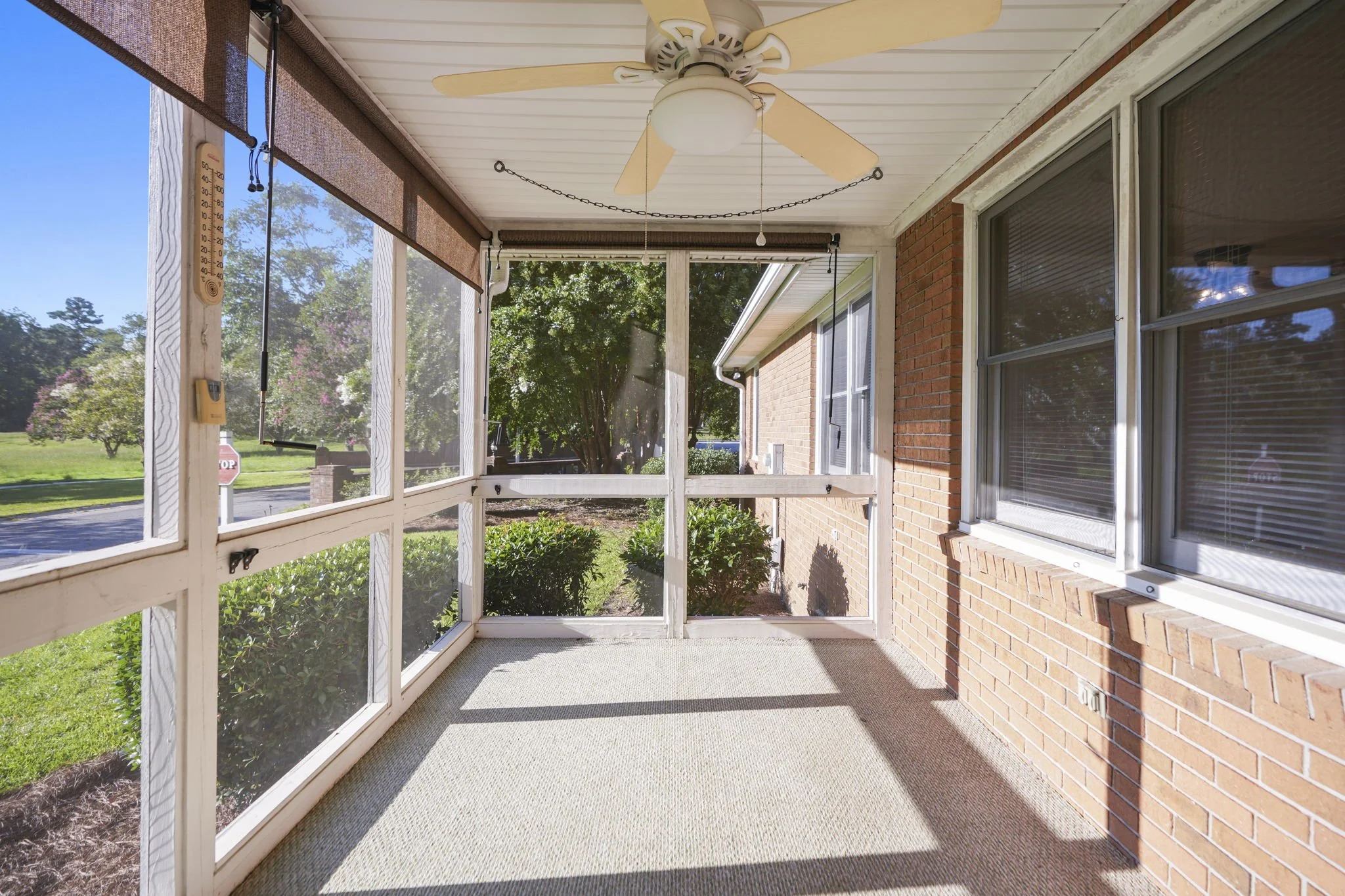 Enclosed porch with ceiling fan, hanging thermometers, and large windows showing landscaped yard with bushes and trees.