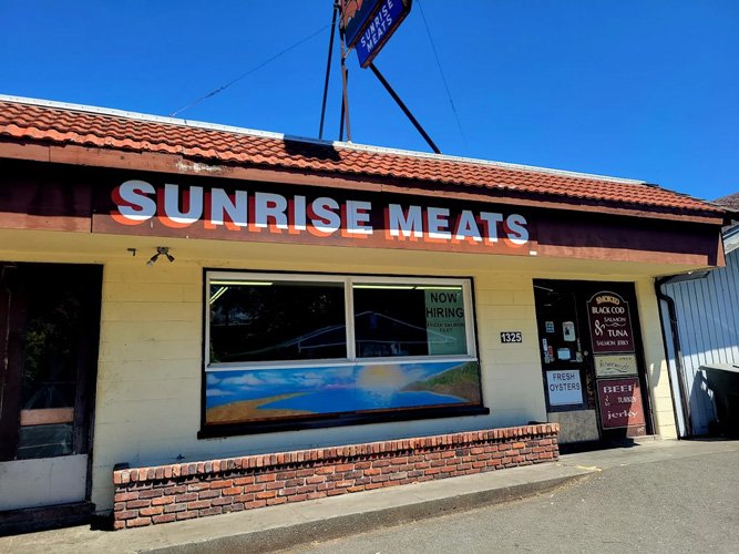 Exterior view of Sunrise Meats, a small meat shop with a red tile roof and cream-colored walls, featuring a large window, a sign indicating they are hiring, and signs for smoked salmon, tuna, fresh oysters, beef, and jerky.