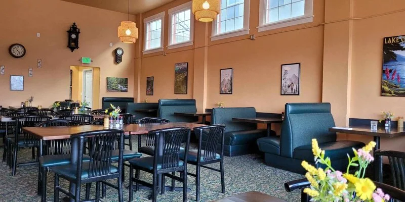 Interior of a dining area with wooden tables, black chairs, teal booth seating, framed pictures on the wall, and large windows letting in natural light.
