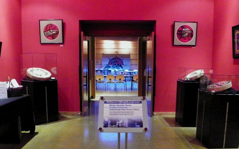 Room with pink walls, framed circular art, display cases with phones, and a sign at the entrance to a gallery or event space.