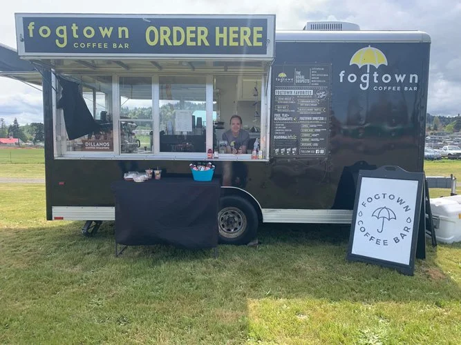 Black coffee truck with a yellow umbrella logo labeled 'Fogtown Coffee Bar' and an 'Order Here' sign on top. A young woman is inside, and a menu is displayed on the side of the truck. There's a small table with a black cover in front of the truck and a sandwich board sign on the grass advertising 'Fogtown Coffee Bar' with an umbrella graphic.