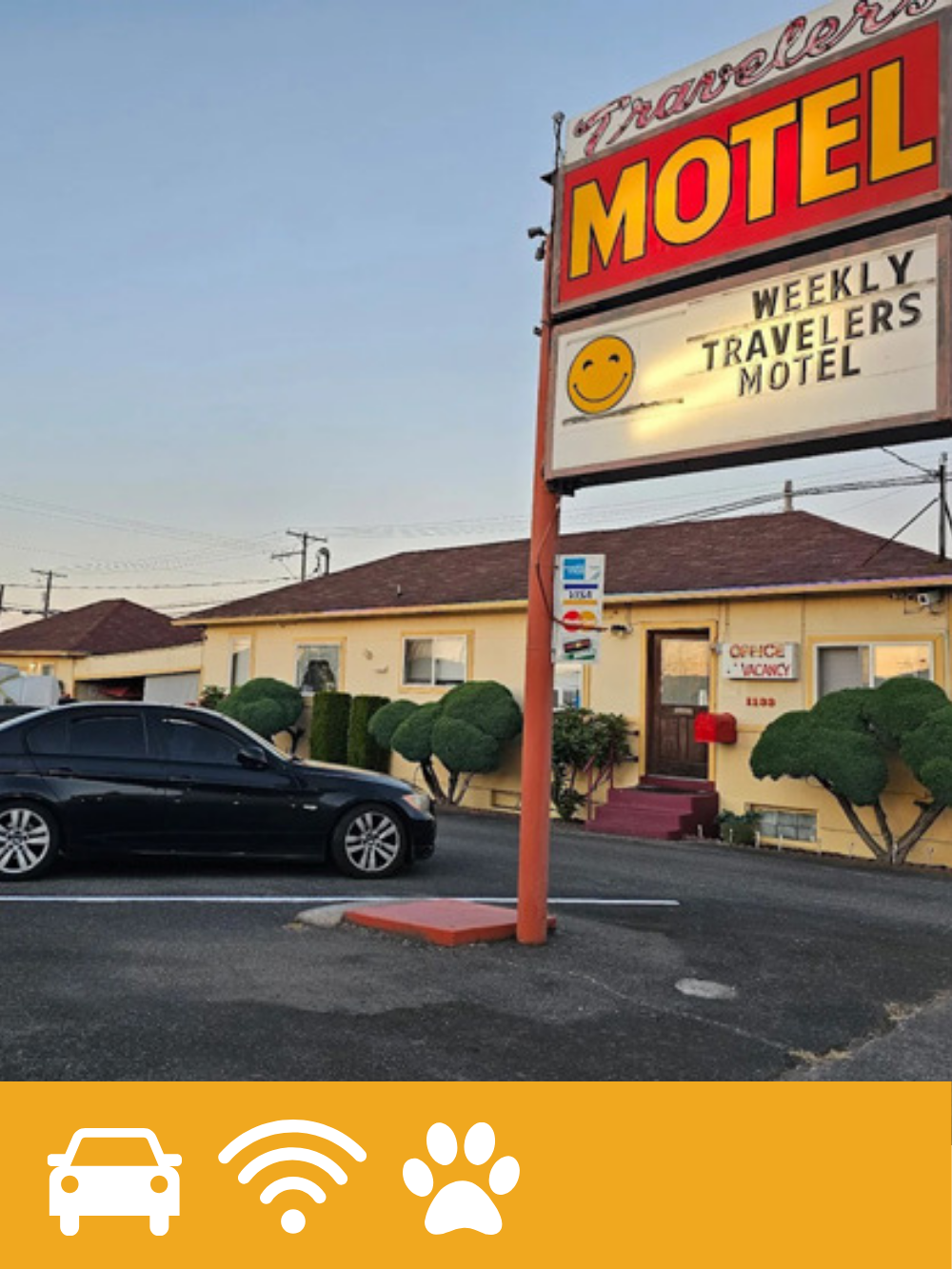 A vintage roadside motel sign advertising Travelers Motel with a smiley face, positioned in front of a yellow single-story building with landscaping and a parking lot with a black car.