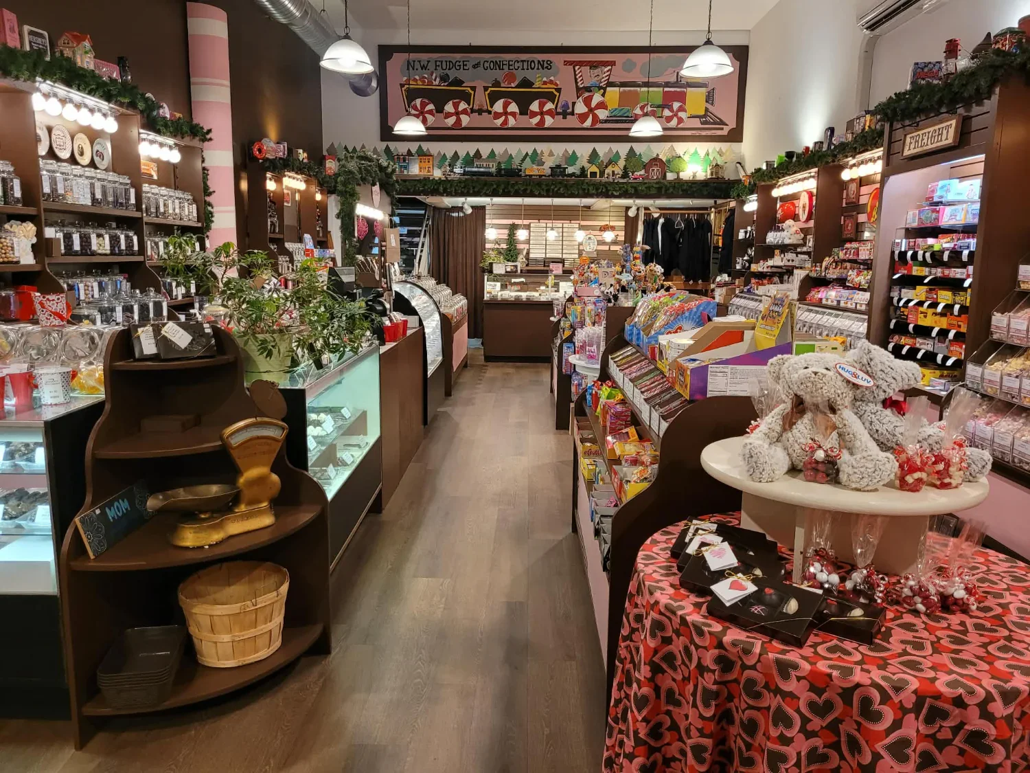 A display case filled with various types of fudge for sale, with a bright red and white banner reading "FRESH FUDGE" hanging below. Behind the case, there are signs and labels, a scale, and shelves with additional items.