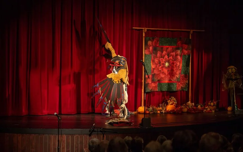 A performer in a colorful costume on stage with red curtains, a large floral quilt backdrop, and pumpkins and flowers on the floor, during a cultural or theatrical event.