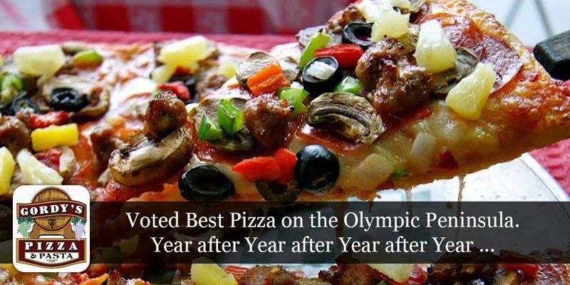 A slice of pizza topped with black olives, green onions, mushrooms, pineapple chunks, and sausage, with more pizza in the background.