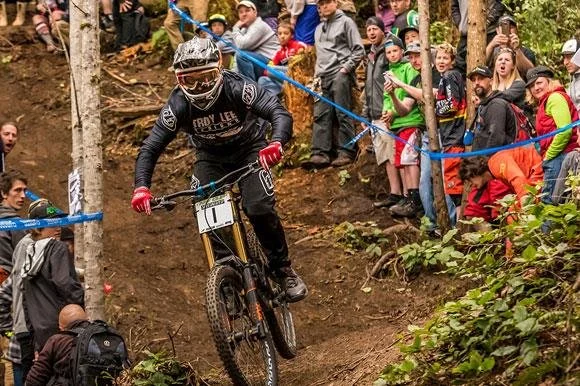 A mountain biker in a black and gold outfit, wearing a helmet and goggles, racing downhill on a dirt trail surrounded by spectators in a wooded area.