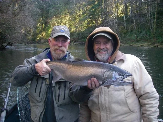 Two men in jackets and hats holding a large fish on a river, with trees in the background.