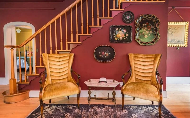 Living room with two vintage yellow upholstered armchairs, a small marble-top table in between, and a staircase with wooden spindles against a burgundy wall decorated with floral paintings and a framed picture.