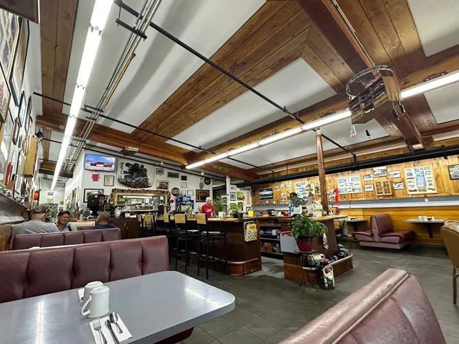 Inside a restaurant with booths, tables, and a bar area, decorated with pictures and memorabilia on the walls, with several customers and staff present.