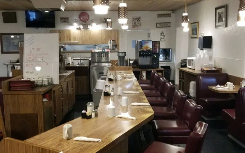 Interior of a small restaurant or diner with a wooden counter set for customers, featuring cups, napkins, and condiments, and purple chairs along the counter. The background includes a whiteboard, kitchen area with appliances, and framed pictures on the walls.