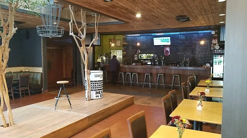 Interior of a cozy cafe or restaurant with wooden furniture, decorated with small flower vases on tables, and a stage with a bar counter in the background.