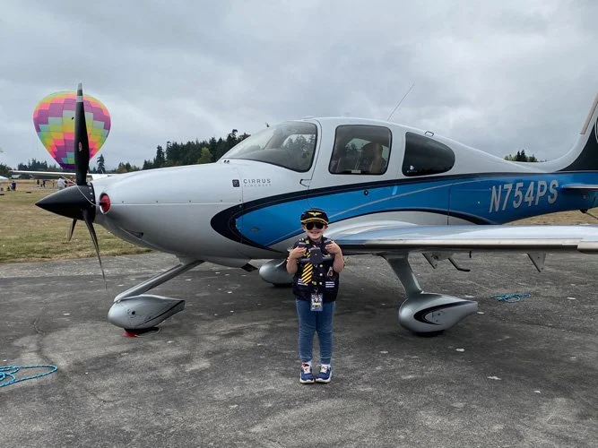 A young girl standing in front of a small private airplane on an airstrip, with a colorful hot air balloon in the background.